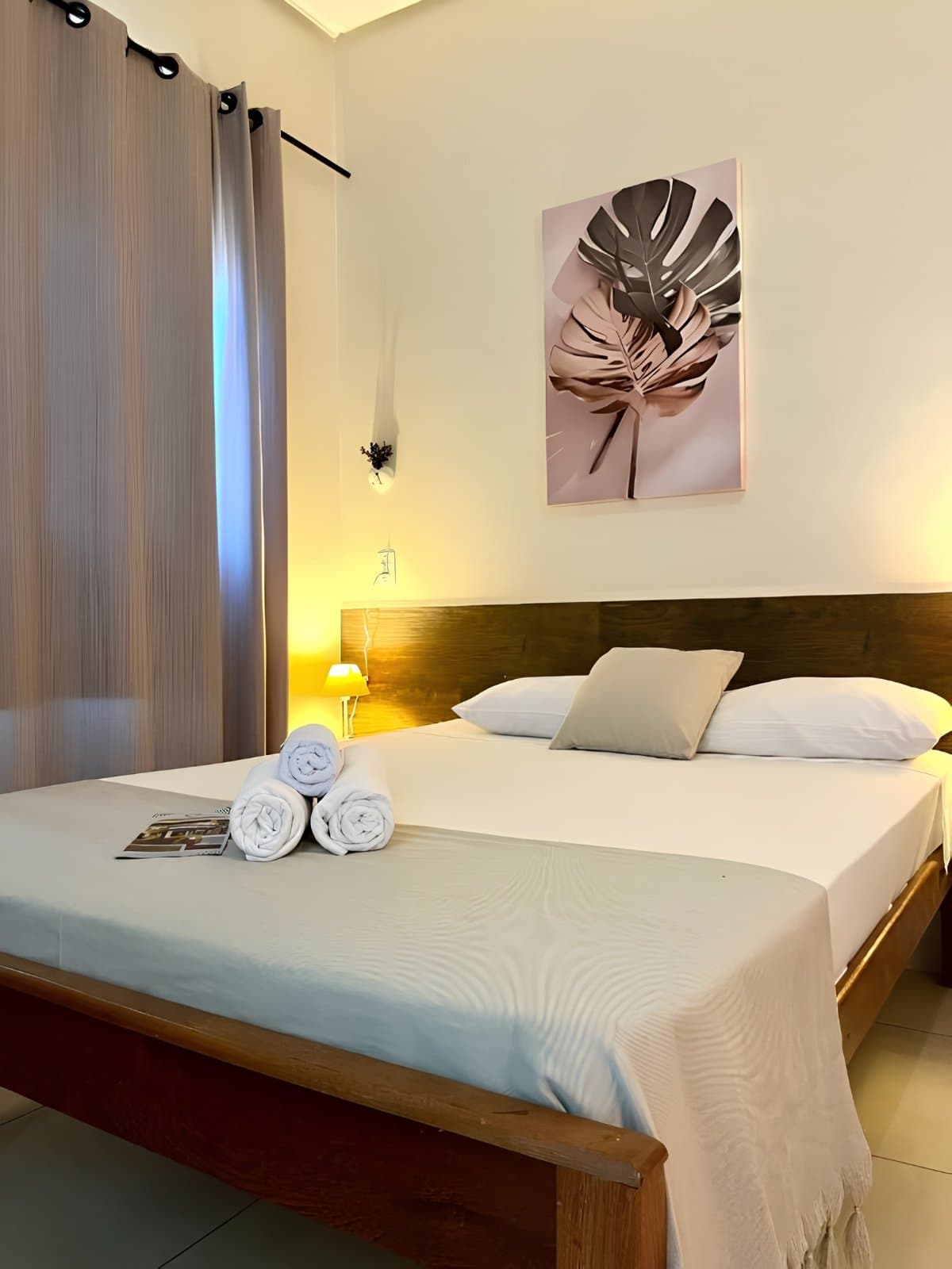 Cama feita, luz quente, e o descanso de quem passou o dia na trilha. Cama de casal arrumada com toalhas brancas enroladas e quadro botânico na parede de um quarto aconchegante de pousada em uma aventura no Jalapão