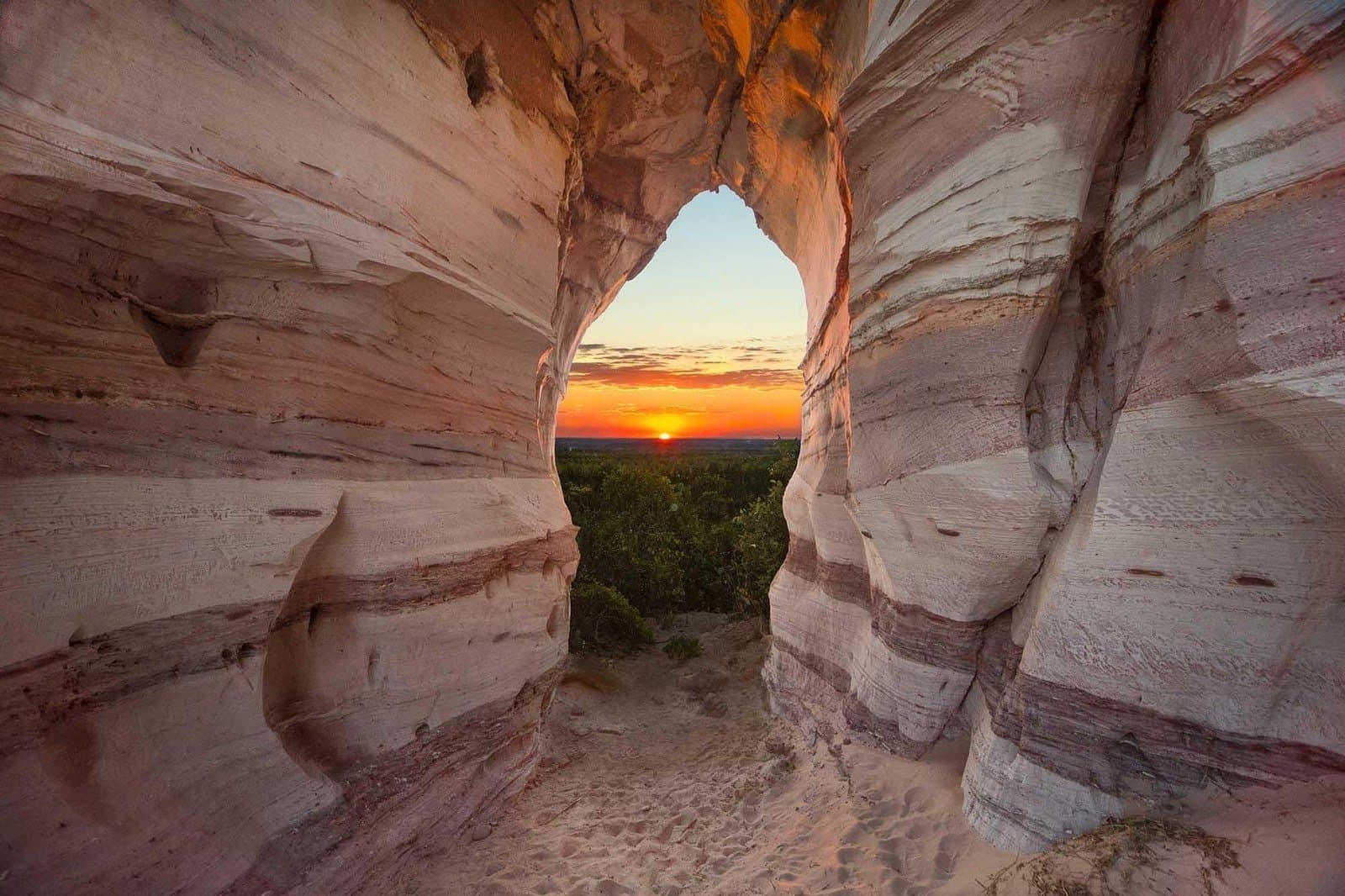 Rocha antiga, um buraco certeiro, e o sol resolveu aparecer com tudo. Vista através de um arco natural de arenito na Pedra Furada, emoldurando um pôr do sol alaranjado sobre o cerrado verde durante uma aventura em Jalapão