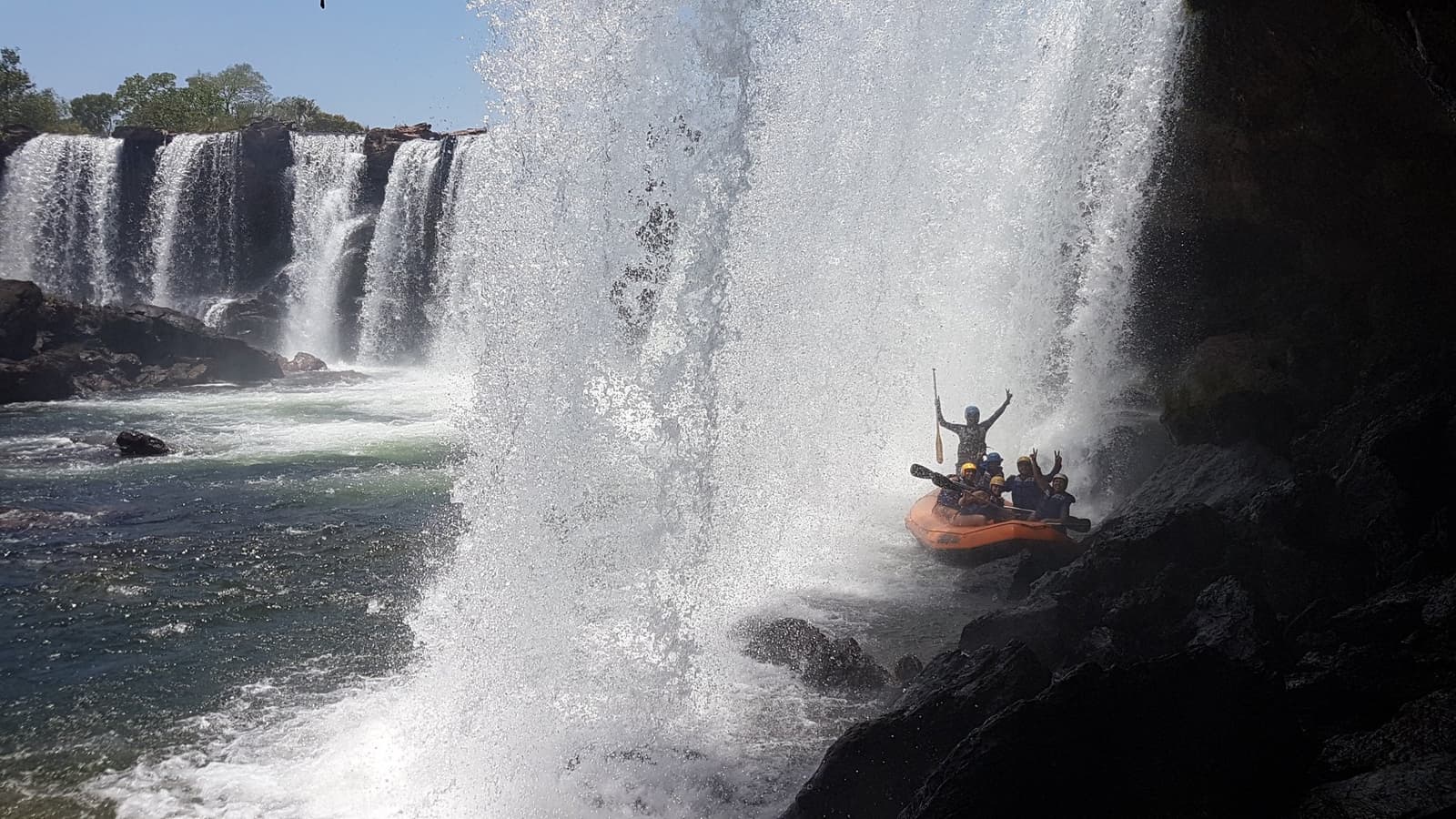 Remo erguido, todo mundo encharcado, e o sorriso que não sai. Um grupo de participantes com capacetes em um bote laranja ergue os braços sob a cortina de água de uma cachoeira larga durante uma aventura no Jalapão, com névoa e água branca em volta