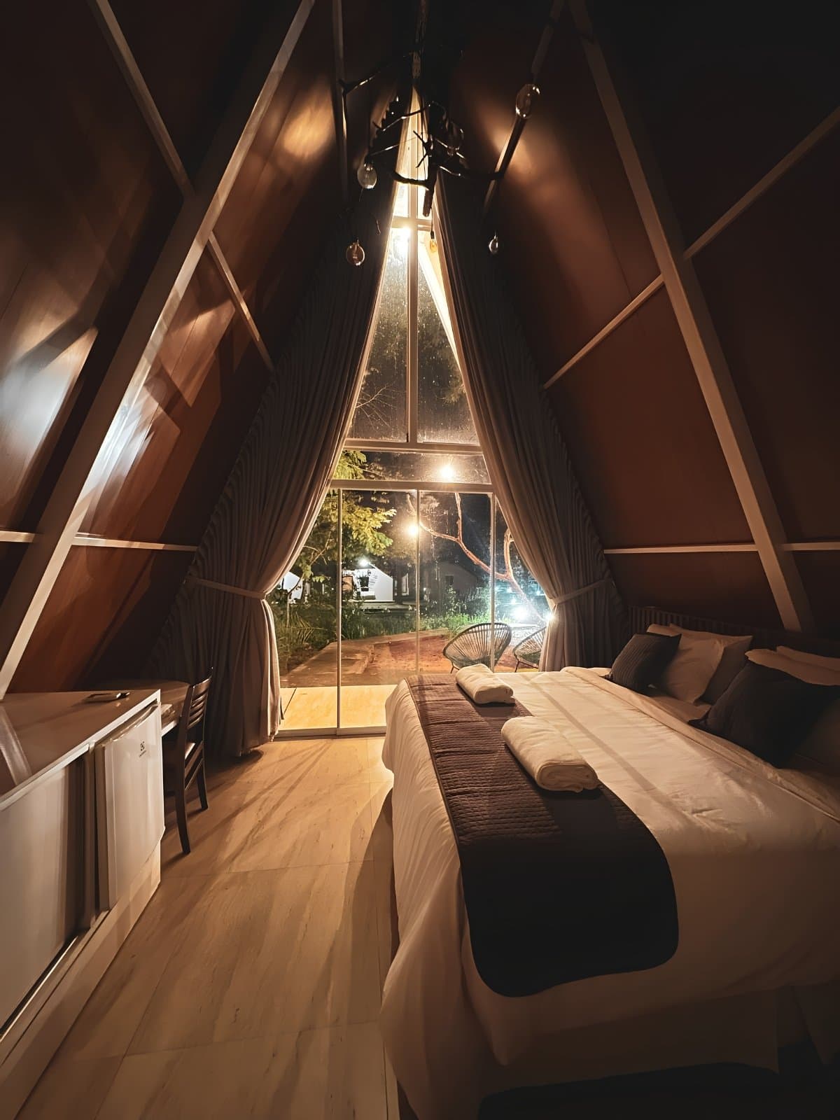 Nach einem langen Tag auf Schotterpisten schläft man hier erstaunlich gut. Inneres einer hölzernen A-Rahmen-Hütte bei Nacht auf einer Jalapão-Reise, mit einem Doppelbett in weißer Bettwäsche, einem hohen dreieckigen Glasfenster mit Blick auf einen beleuchteten Garten und warmem Glühbirnenlicht