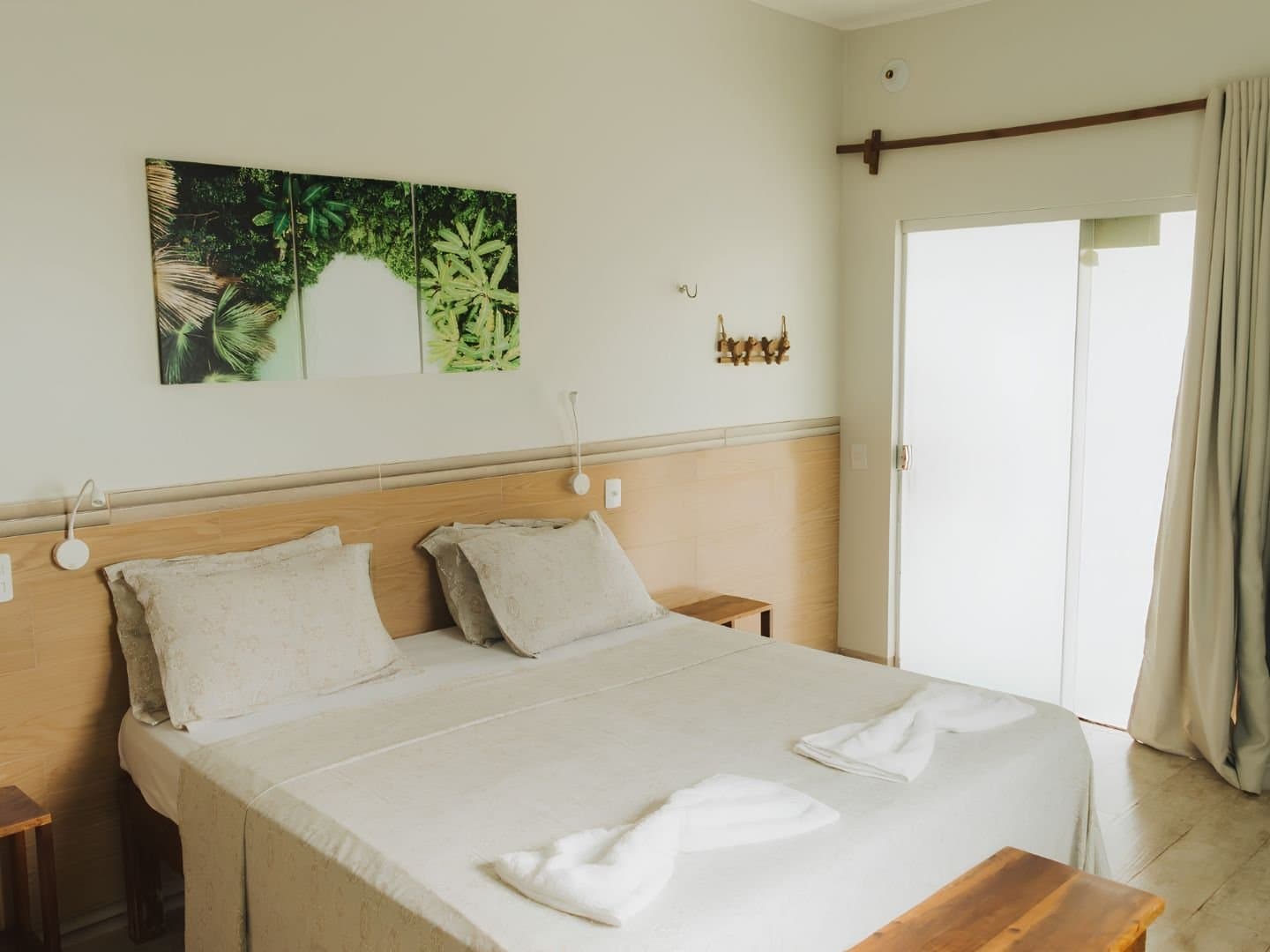 Einfach, sauber und genau das Richtige nach einem langen Tag auf dem Trail. Helles Doppelzimmer einer Jalapão-Unterkunft mit Holzkopfteil, frisch bezogenem weißen Bett, gefalteten Handtüchern, tropischen Naturbildern und einer Schiebetür zum Tageslicht