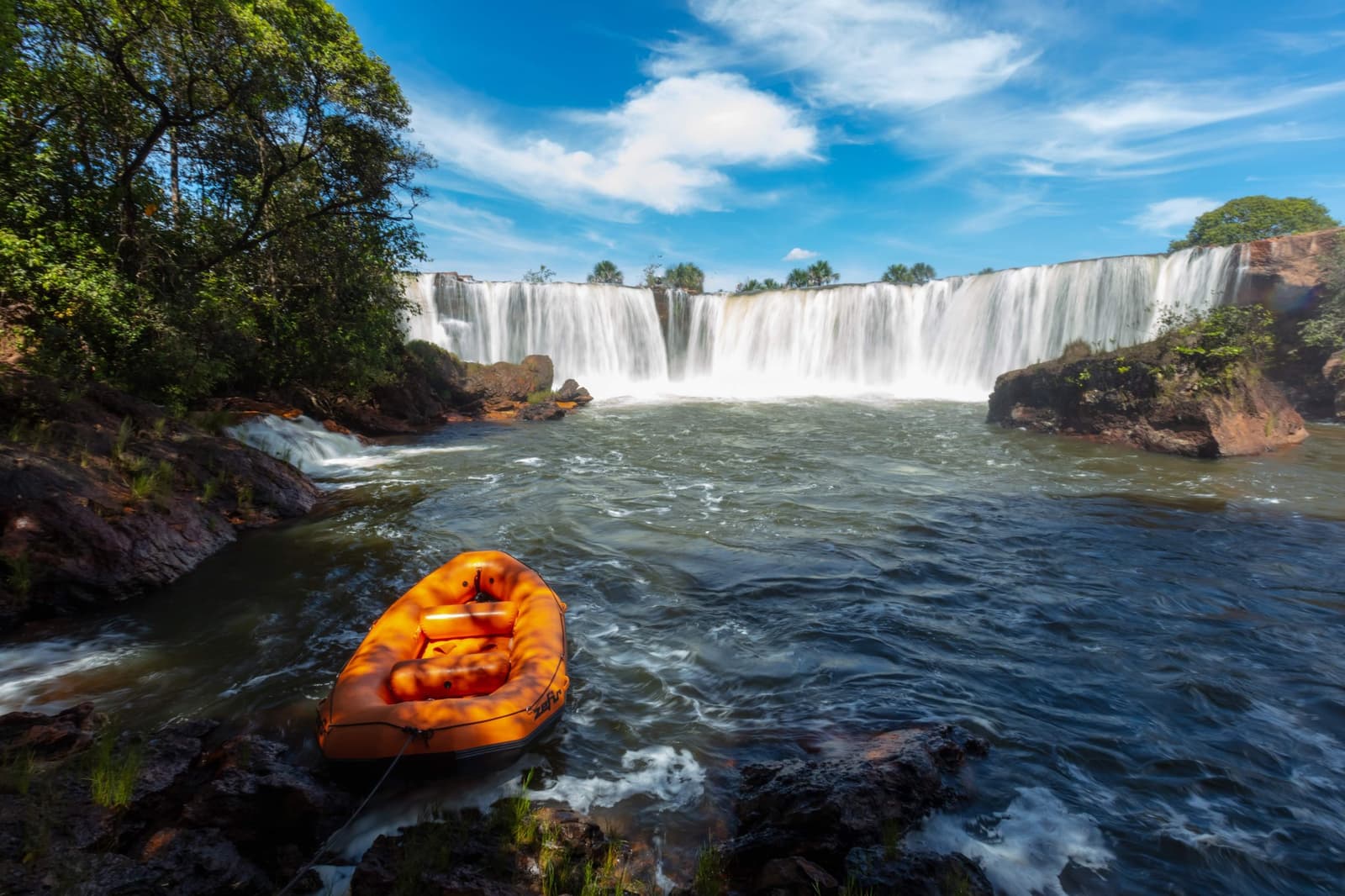 Das Boot wartet geduldig. Der Wasserfall nicht. Photo: Alberto Andrich Ein orangefarbenes Schlauchboot liegt am Felsenufer vor einem breiten Wasserfall auf einer Jalapão-Reise, umgeben von grüner Cerrado-Vegetation unter blauem Himmel
