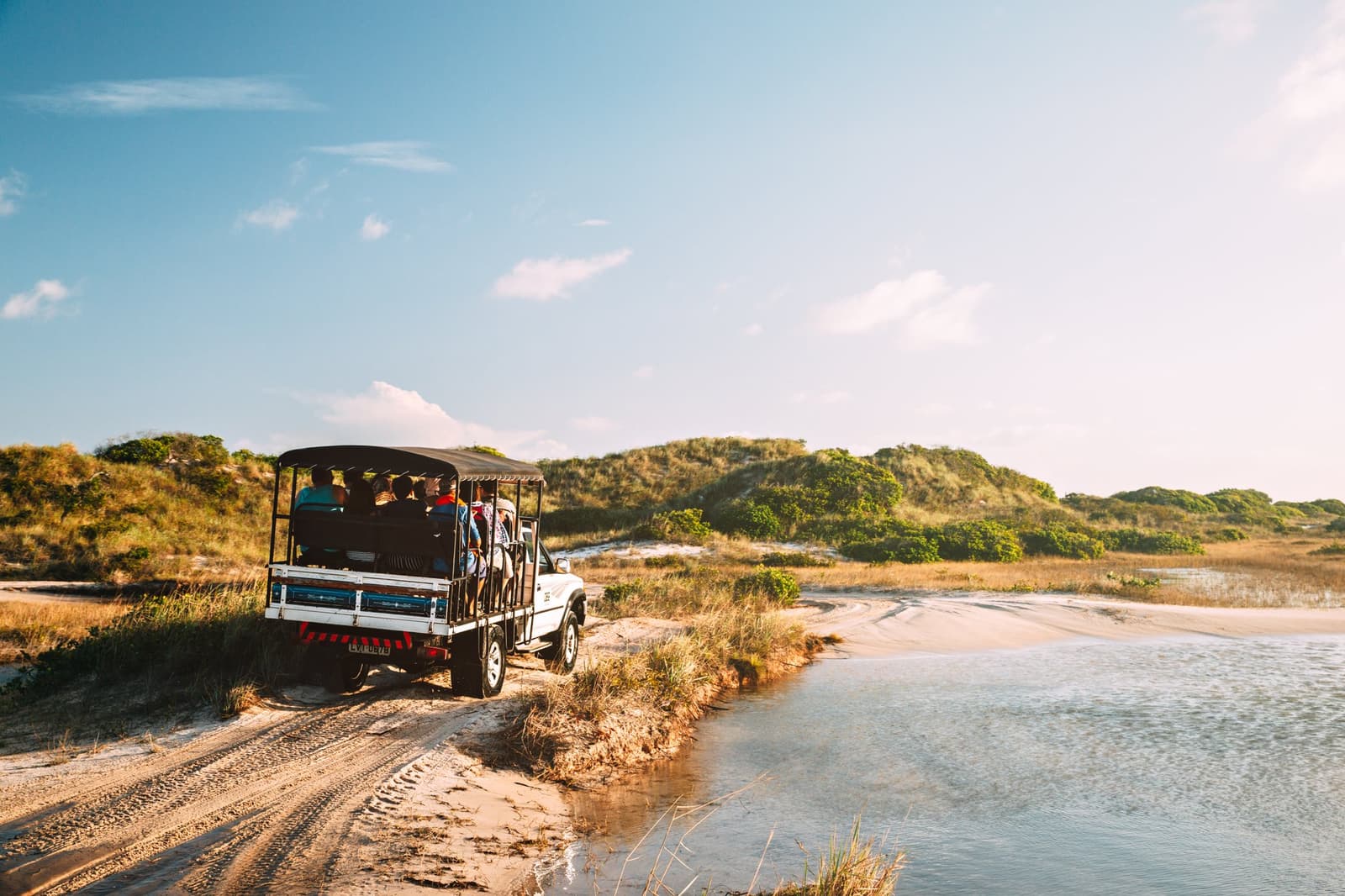 Sandpiste, Lagune, blaues Licht, Platz auf der Ladefläche ist der beste im ganzen Bus. Ein offener 4x4-Geländewagen mit einer Reisegruppe fährt auf einem Sandweg entlang einer flachen Küstenlagune, umgeben von begrünten Dünen und blauem Himmel beim Lençóis Maranhenses Full Itinerary