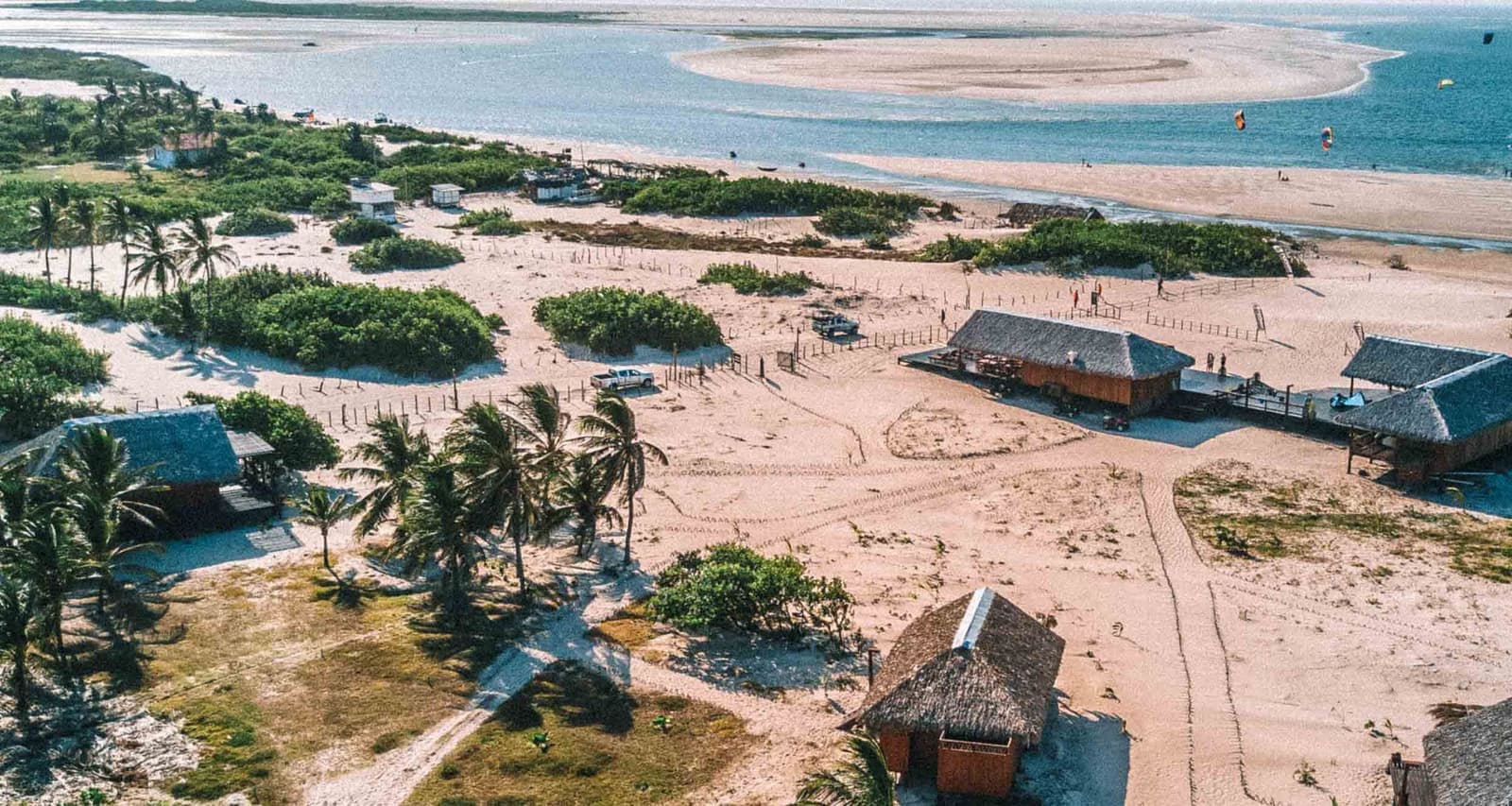 Ein Dorf, das sich zwischen Sand und Meer eingefügt hat, als wäre es nie anders gewesen. Luftaufnahme von strohgedeckten Hütten, Palmen und Sandwegen im rustikalen Dorf Atins mit Kitesurfern auf dem blauen Wasser im Hintergrund