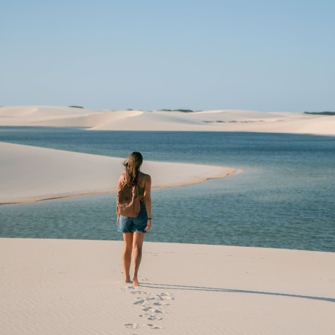Sanddünen, Süßwasserlagunen und kein Ziel außer dem nächsten Schritt. Eine Frau mit Rucksack läuft barfuß über weiße Sanddünen auf eine kristallklare blaue Lagune in den Lençóis Maranhenses zu, ihre Fußspuren im weichen Sand hinter ihr