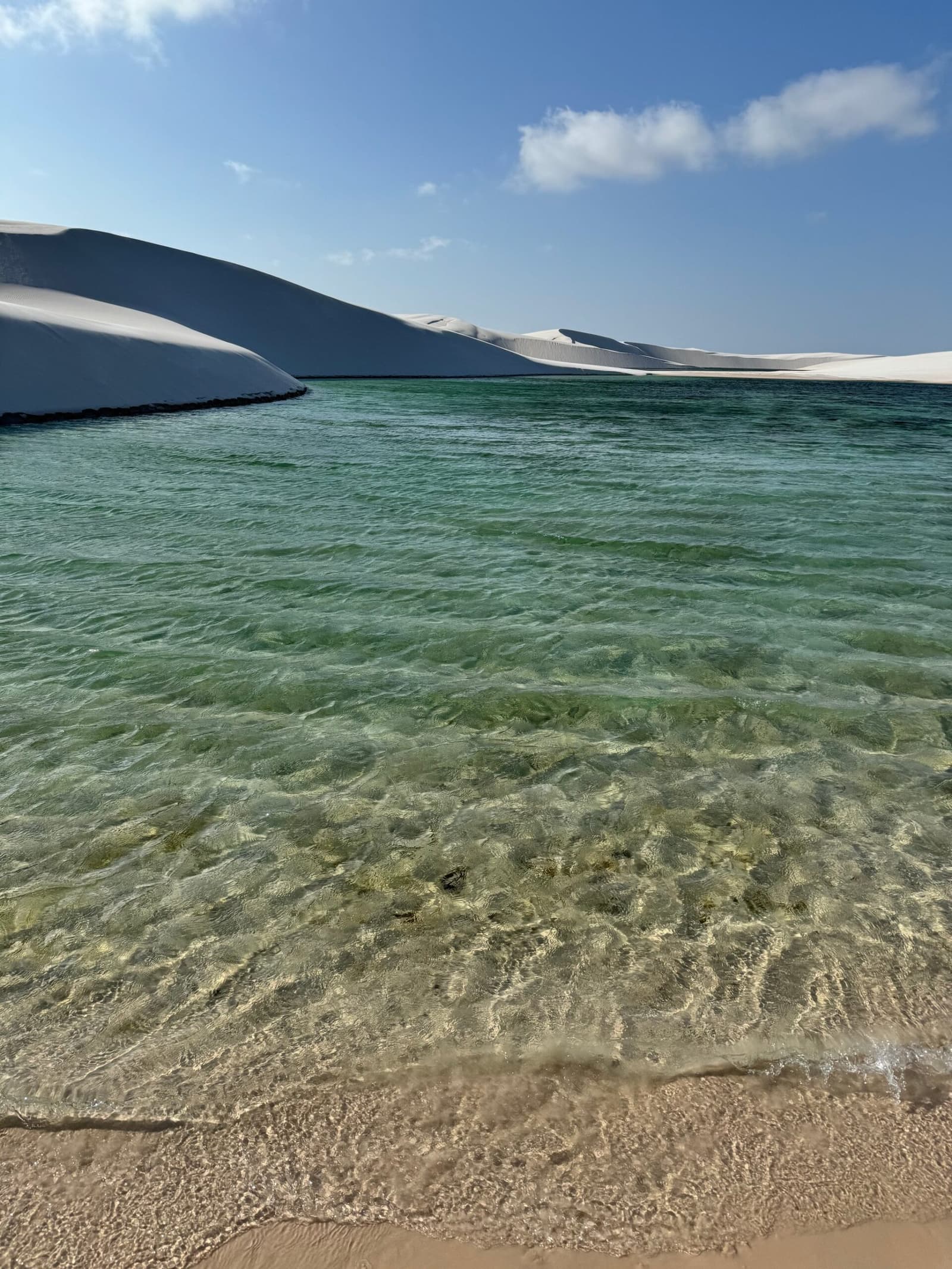 Kühl, klar und nach der Wanderung durch die Dünen genau das Richtige. Kristallklares grünes Wasser einer Süßwasserlagune am Sandstrand beim Lençóis-Maranhenses-Hike, mit hohen weißen Sanddünen entlang des gegenüberliegenden Ufers unter blauem Himmel