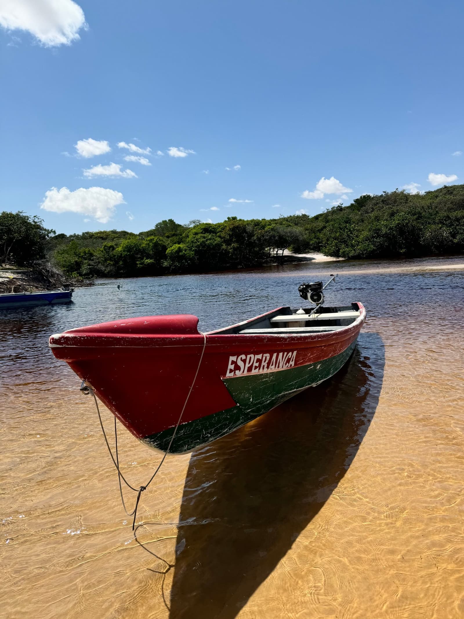 Hoffnung liegt hier direkt am Ufer, bereit zum Ablegen. Ein rot-grünes Holzboot namens Esperança liegt am flachen Sandufer eines dunklen Flusses, mit einer weißen Sandbank und grüner Vegetation am gegenüberliegenden Ufer unter blauem Himmel