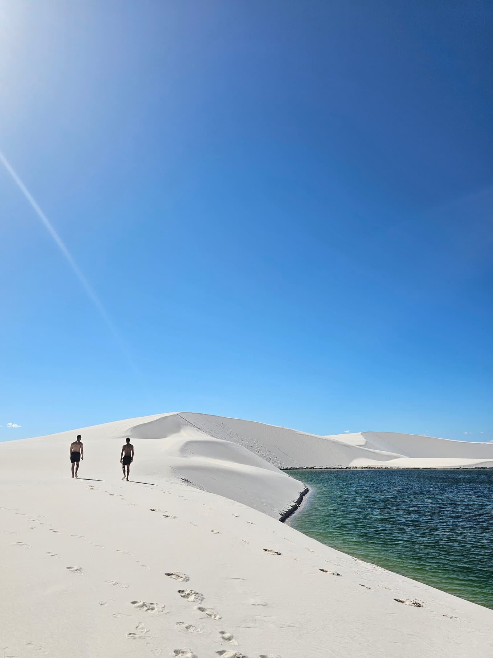 Weißer Sand, smaragdgrünes Wasser und das gute Gefühl, genau dort zu sein. Zwei Wanderer gehen barfuß eine riesige weiße Sanddüne neben einer klaren grünen Lagune entlang, beim Wandern in den Lençóis Maranhenses, unter einem tiefblauen Mittagshimmel