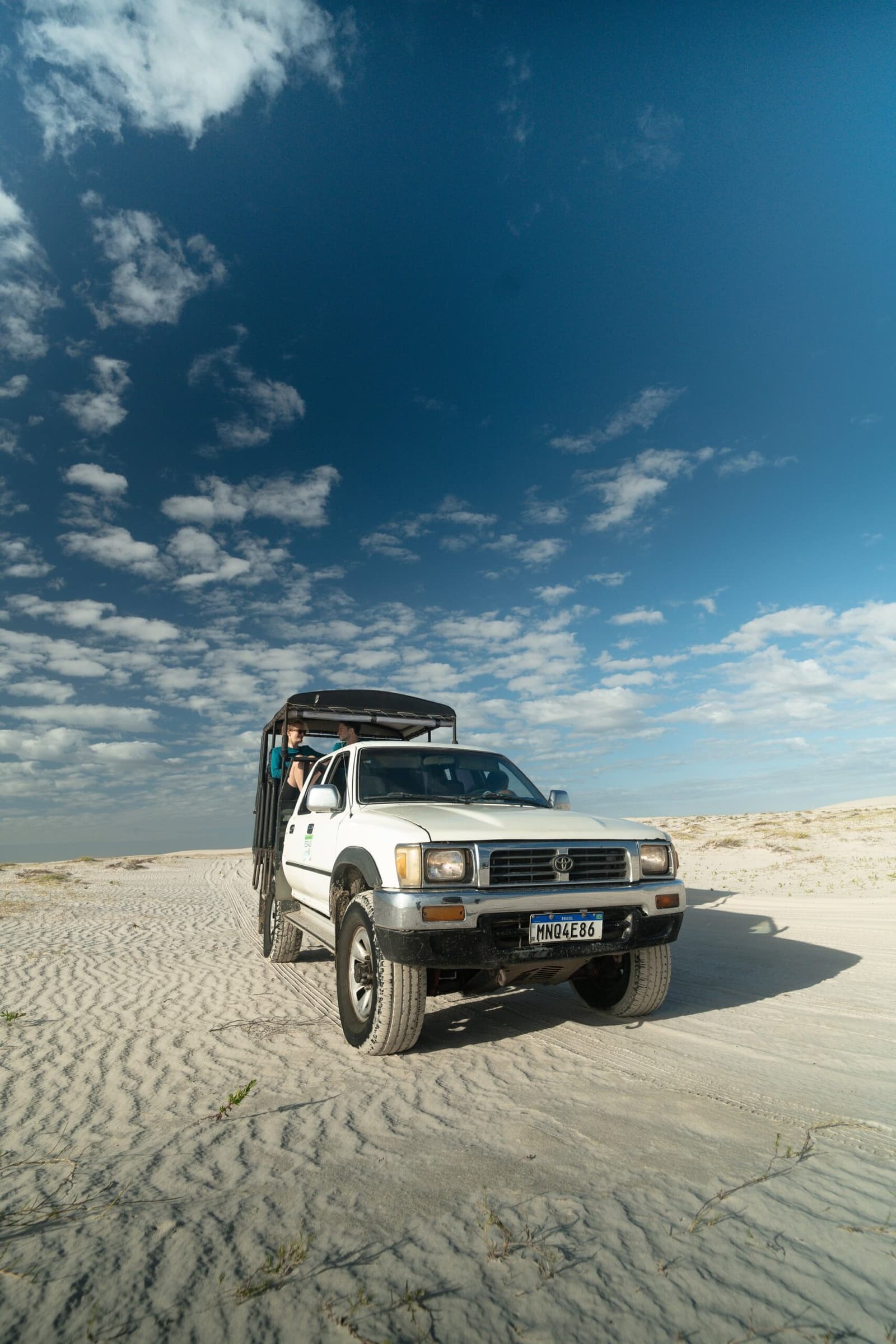 Le vrai luxe ici, c'est le ciel au-dessus de la benne. Un Toyota 4x4 blanc avec une benne ouverte et des passagers debout traverse de vastes dunes de sable blanc sous un ciel bleu parsemé de nuages, au début d'un trek dans les Lençóis Maranhenses