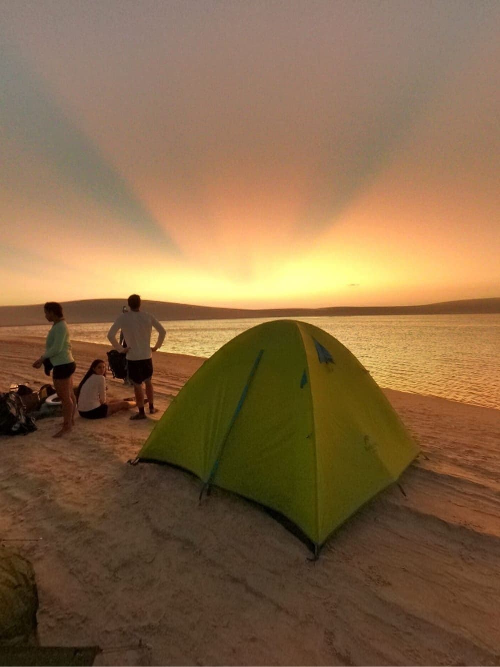 Le sable encore chaud, le lagon qui s'embrase, et personne ne presse. Une tente dôme verte installée sur une plage de sable lors d'un trek dans les Lençóis Maranhenses, avec trois voyageurs contemplant un coucher de soleil doré intense sur une eau calme et des dunes au loin