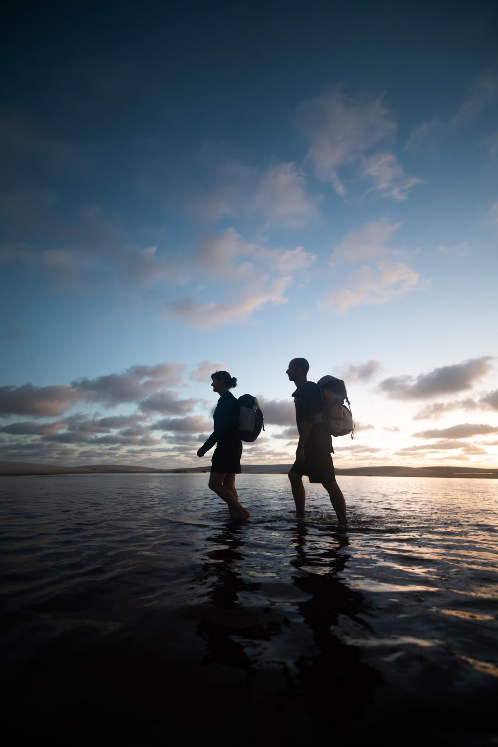 Les pieds dans l'eau, le ciel pour soi, et l'heure qui n'a plus d'importance. Deux randonneurs avec sacs à dos en silhouette contre un ciel crépusculaire, marchant pieds nus dans une eau peu profonde à la fin d'une journée de trekking aux Lençóis Maranhenses