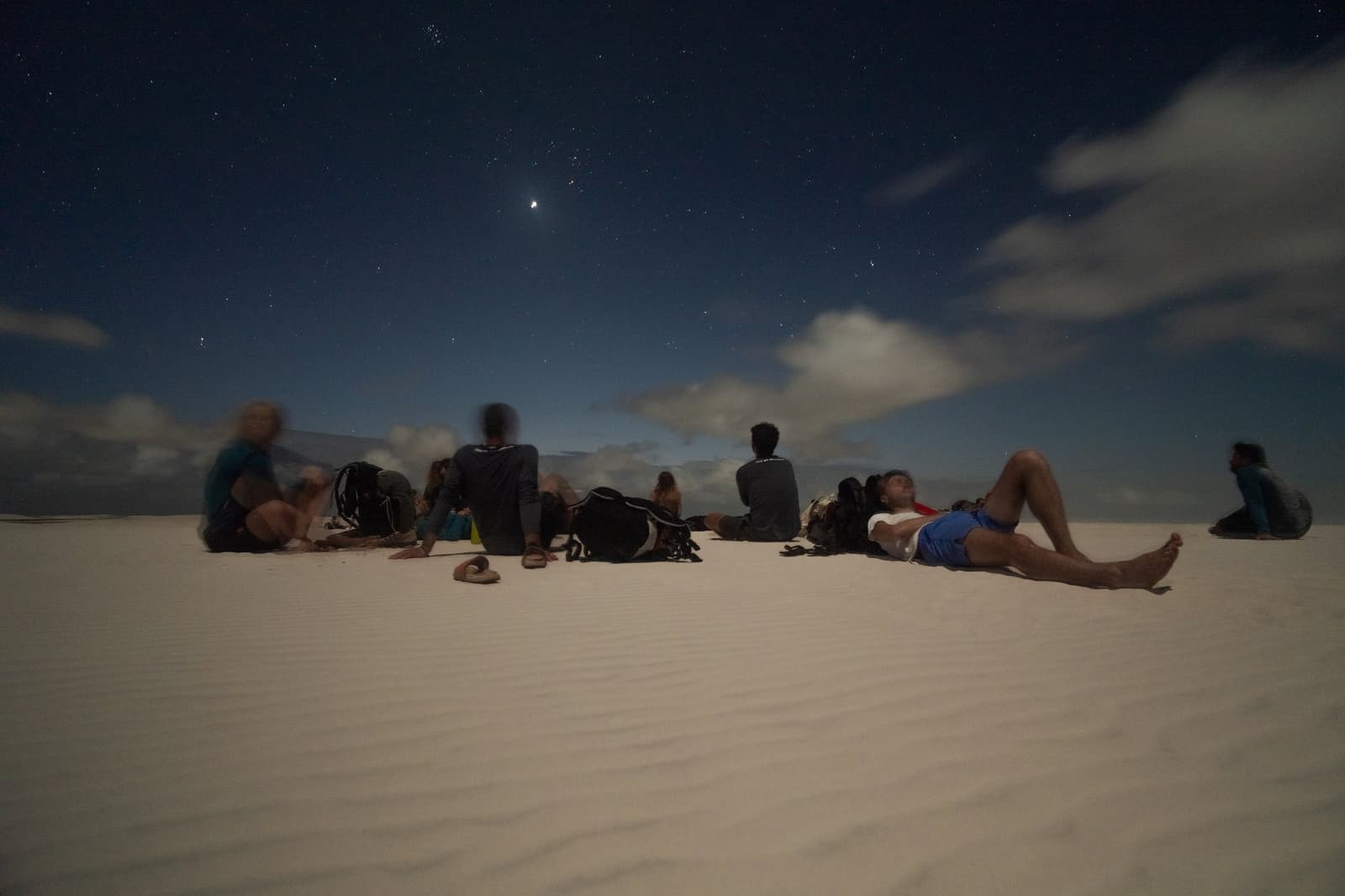 Pas de transats ici, juste du sable chaud, des étoiles et de belles rencontres. Un groupe de voyageurs allongés et assis sur les dunes de sable blanc la nuit lors d'un trek dans les Lençóis Maranhenses, contemplant un ciel étoilé parsemé de nuages avec une planète brillante visible au-dessus
