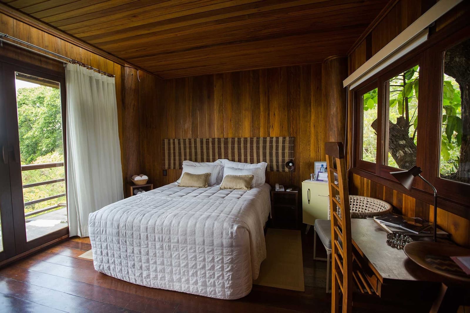 Einschlafen mit dem Dschungel direkt vor dem Fenster. Ein Gästezimmer im Mirante do Gavião mit warmen Holzwänden, Decke und Boden, einem weißen gesteppten Doppelbett, gewebtem Kopfteil, Holzschreibtisch und Fenstern mit Blick auf üppige Dschungelvegetation
