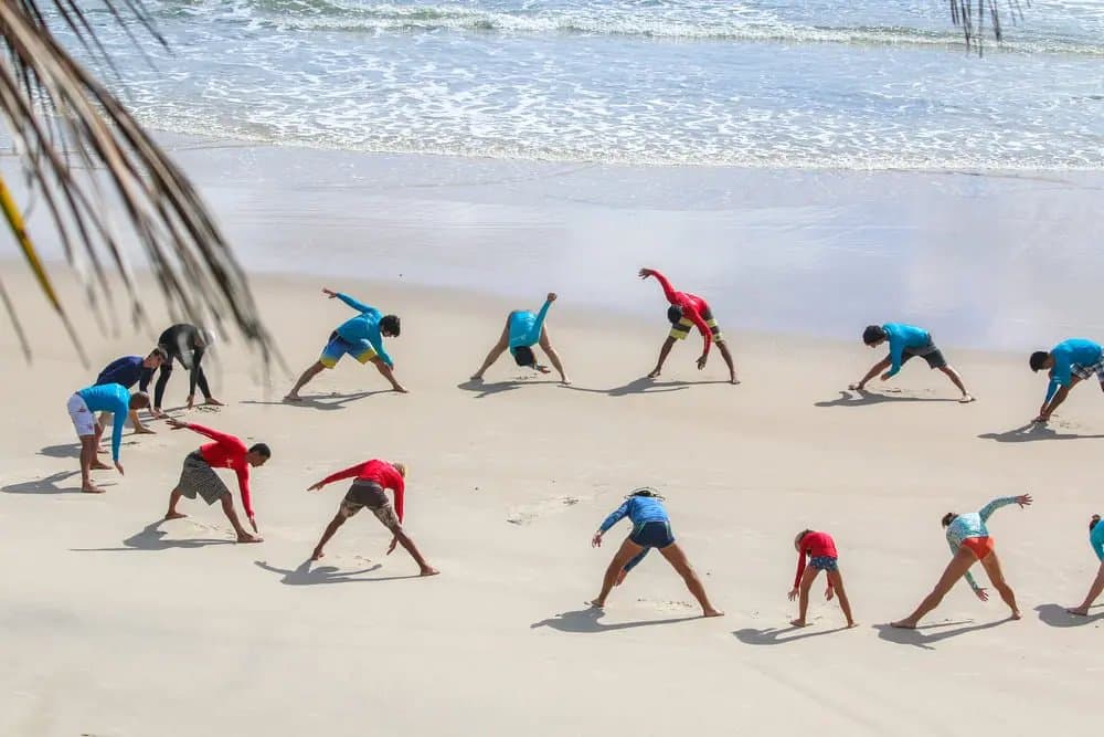 O aquecimento que separa os animados dos muito animados. Um grupo de alunos com lycras faz exercícios de alongamento em uma praia de areia branca antes das aulas de surf em Itacaré, com ondas suaves do oceano ao fundo e uma folha de palmeira em primeiro plano.