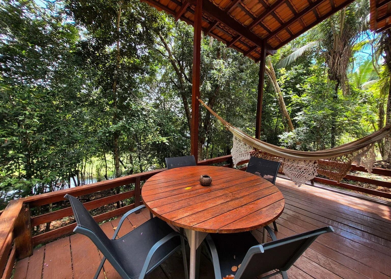 Mittagessen, Hängematte, Wald, mehr braucht es eigentlich nicht. Überdachte Holzterrasse einer Amazon-Boutique-Lodge mit rundem Holztisch, vier Stühlen, zwei Hängematten aus Makramee und einem dichten Regenwalddach sowie einem Wasserlauf hinter dem Geländer
