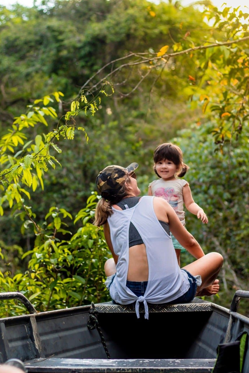 Wenn Kinder dabei sind, wird selbst die Anfahrt zum Abenteuer. Eine Frau mit Tarnkappe und ein lächelndes Kleinkind sitzen gemeinsam auf der Ladefläche eines Fahrzeugs, umgeben von üppiger Amazonas-Vegetation während eines Amazon-Boutique-Lodge-Ausflugs