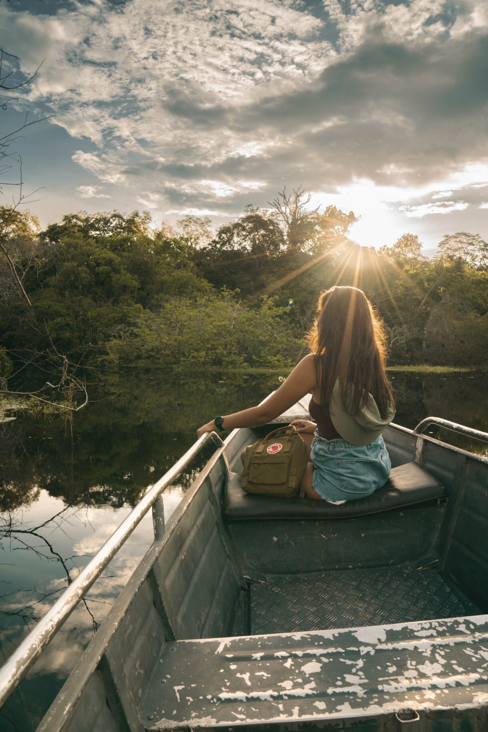 Manche Momente lässt man einfach geschehen, während der Fluss die Zeit übernimmt. Photo: Yuri Panichi Eine Frau sitzt am Bug eines kleinen Aluminiumboots auf einem stillen Fluss und beobachtet, wie die Sonne bei goldener Stunde durch die Baumkronen bricht – ein Ausflug vom Amazon Boutique Lodge