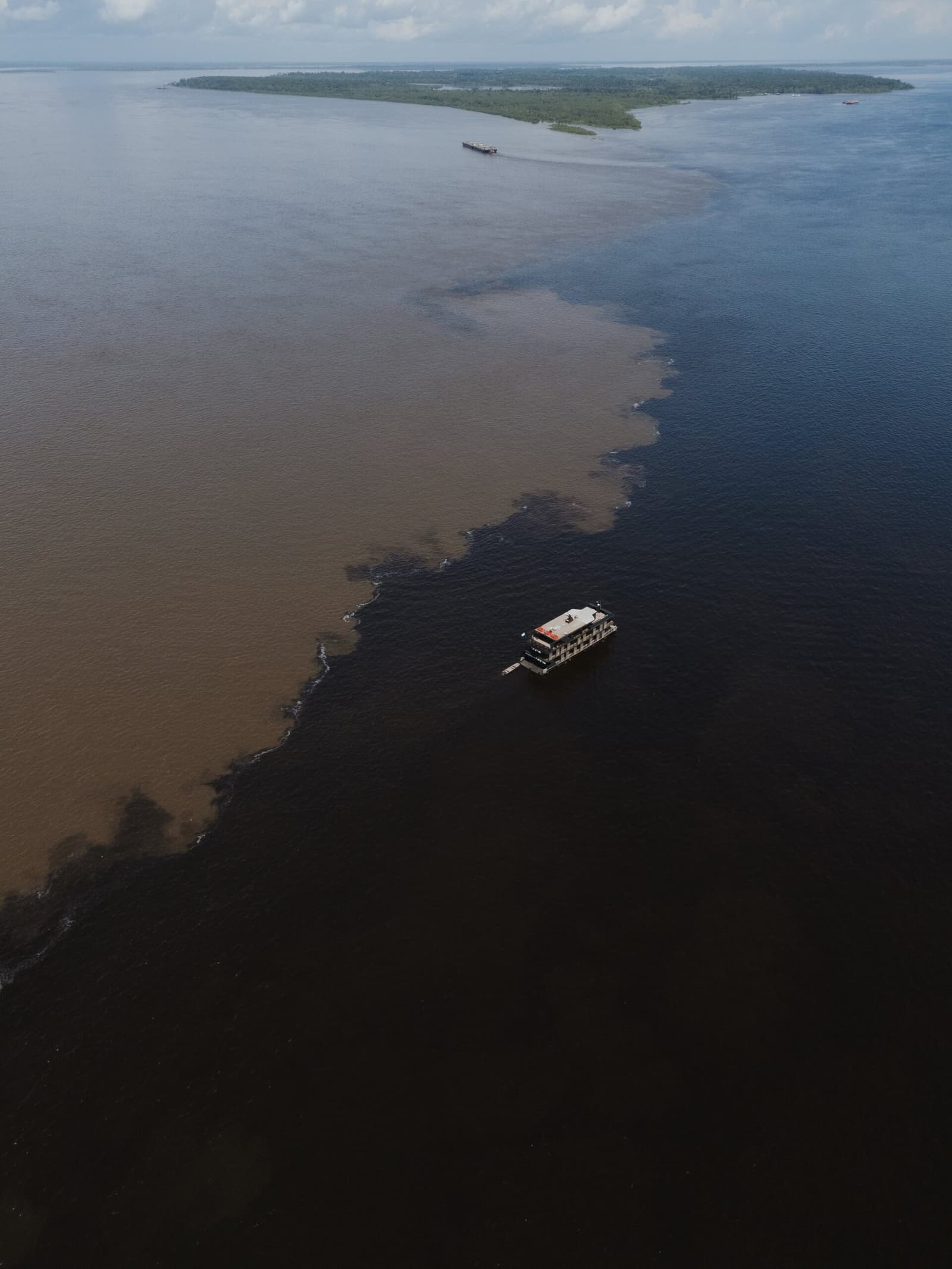 Schwarz trifft Braun, und das Boot fährt genau dazwischen hindurch. Luftaufnahme eines Flussboots, das die Begegnung der Gewässer überquert, wo schwarzes und braunes Wasser in einer auffälligen Grenzlinie aufeinandertreffen, mit einer bewaldeten Insel im Hintergrund