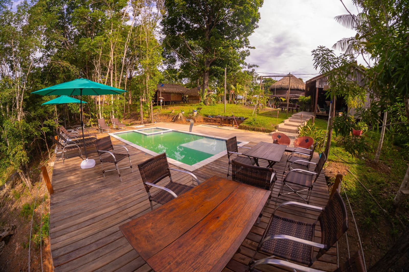 Kühles Wasser, warmes Holz, und dahinter einfach der Dschungel. Holzdeck mit Esstischen, Korbstühlen, türkisfarbenem Pool und Strohdach-Bungalows im Amazon Turtle Lodge, umgeben von hohen Regenwaldäumen