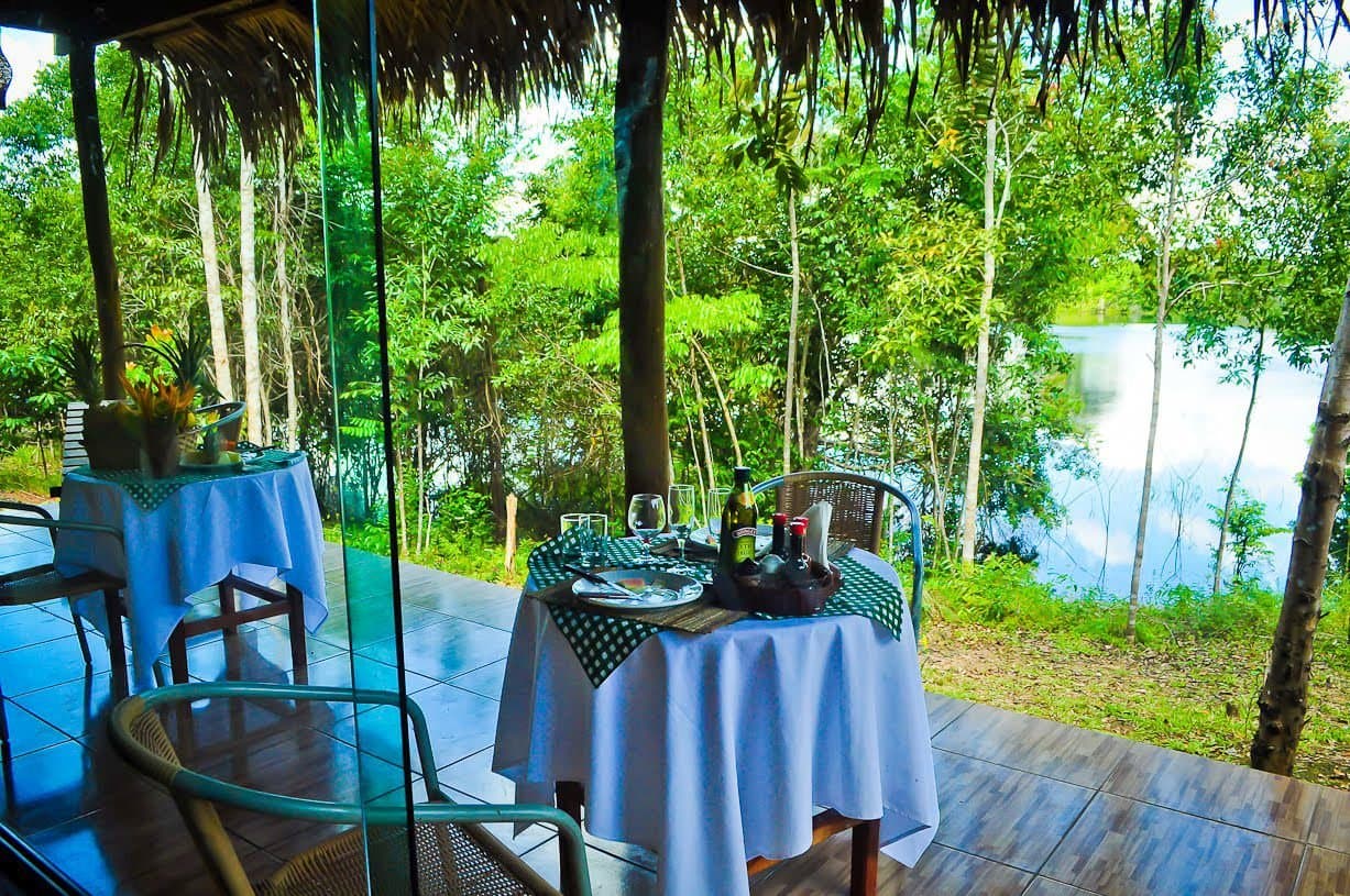 Abendessen, bei dem der Fluss das letzte Wort hat. Überdachte, offene Dining-Terrasse der Amazon Turtle Lodge mit weiß gedeckten Tischen, Weinflaschen und einem ruhigen Fluss inmitten üppiger Regenwaldvegetation
