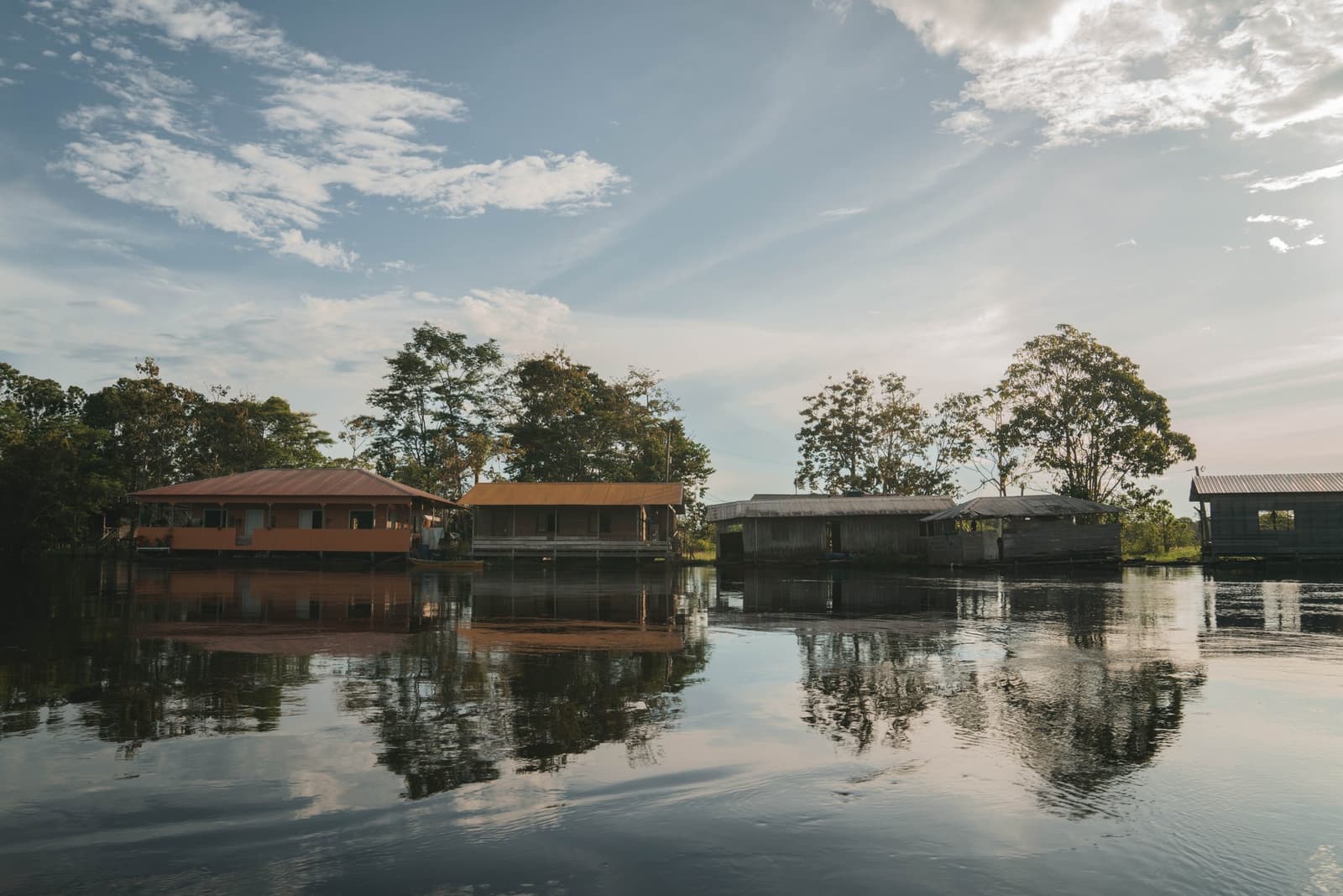 Dein Zuhause für diese Tage, doppelt schön im Fluss gespiegelt. Auf Stelzen gebaute Holz- und gestrichene Gebäude der Amazon Turtle Lodge stehen direkt über einem ruhigen, dunklen Fluss, mit hohen Regenwaldbaümen, die sich im stillen Wasser unter einem teilweise bewölkten Himmel spiegeln