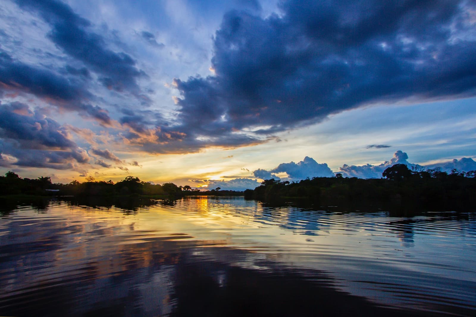 Kein Abend hier gleicht dem vorigen, der Amazonas entscheidet das selbst. Dramatischer Sonnenuntergang über einem breiten Regenwaldflusses, goldenes Licht spiegelt sich auf ruhig plätscherndem Wasser, dunkle Baumsilhouetten am Horizont