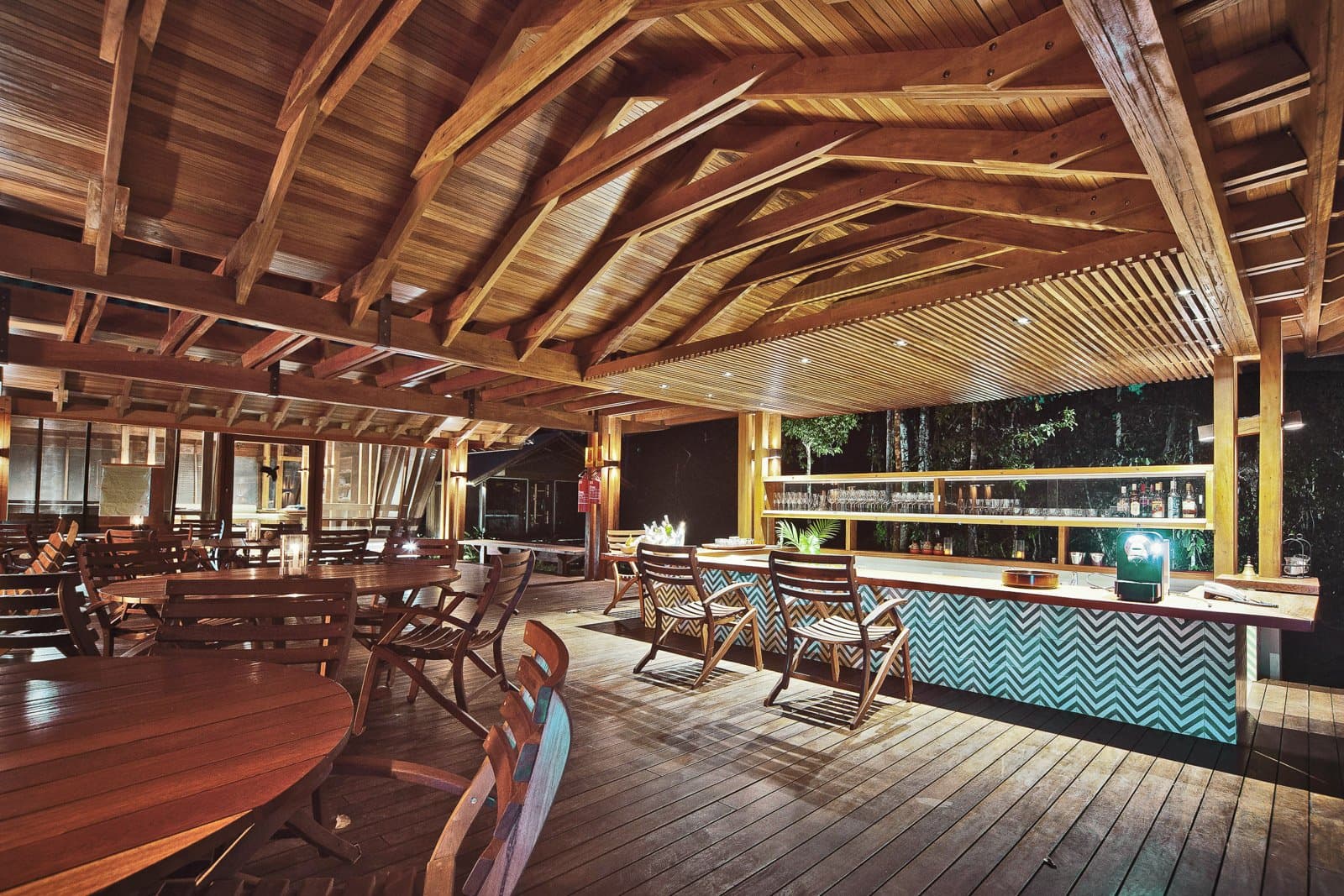 Un verre frais, la forêt qui murmure à deux pas du garde-corps. Photo: Samuel Melim (c) Bar et salle à manger en plein air d'un lodge d'observation des oiseaux en forêt amazonienne, avec des poutres en bois apparentes, un comptoir de bar carrelé en chevrons turquoise, des rangées de verres et de bouteilles, des tables rondes en bois et une vue sur la forêt nocturne