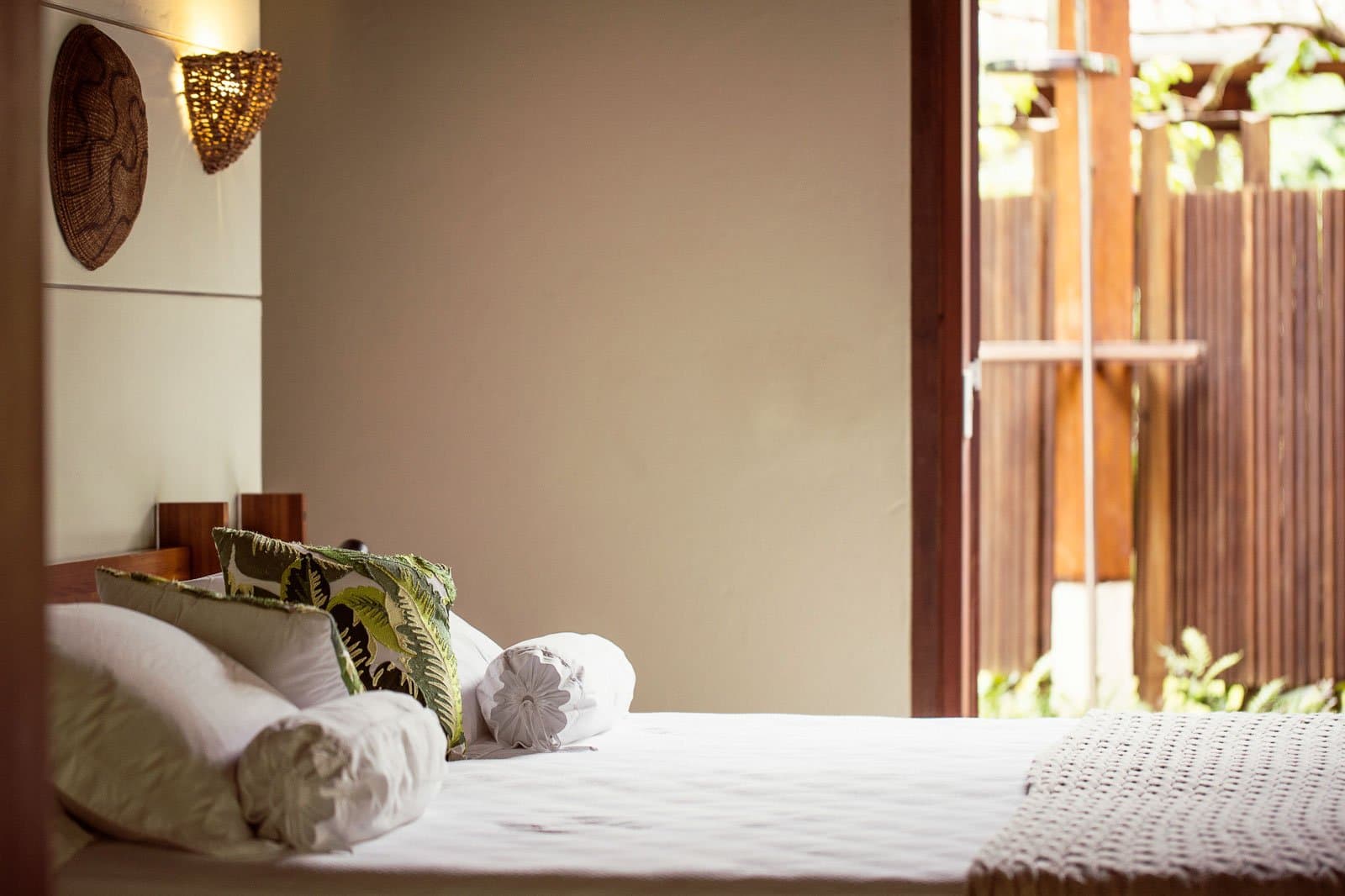 Un vrai lit confortable, parce que le réveil sonne bien avant l'aube. Photo: | SamuelMelim Photography Grand lit soigneusement fait avec des coussins à motifs tropicaux et blancs dans une chambre de lodge d'observation des oiseaux en forêt amazonienne, avec une applique en vannerie tressée et une porte coulissante ouverte sur une terrasse en bois et de la végétation