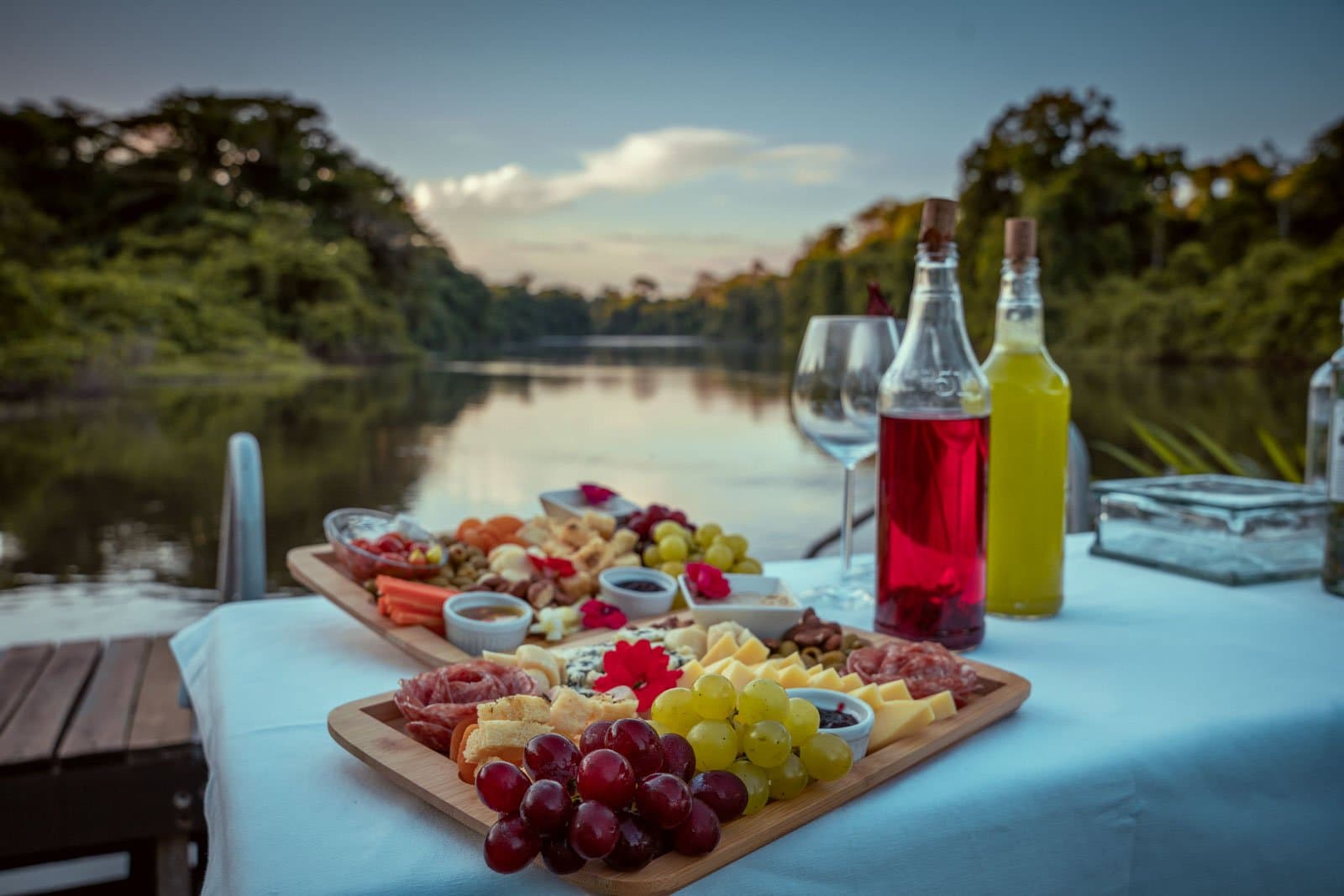 La rivière se tait, le plateau parle pour lui, et la forêt peut patienter encore un peu. Photo: Samuel Melim Un généreux plateau de charcuterie avec raisins, viandes séchées, fromages et sauces disposé sur une nappe blanche au bord d'une rivière calme en pleine jungle au crépuscule, avec deux bouteilles de boissons colorées et des verres à vin, lors d'une expérience d'observation des oiseaux dans la forêt amazonienne
