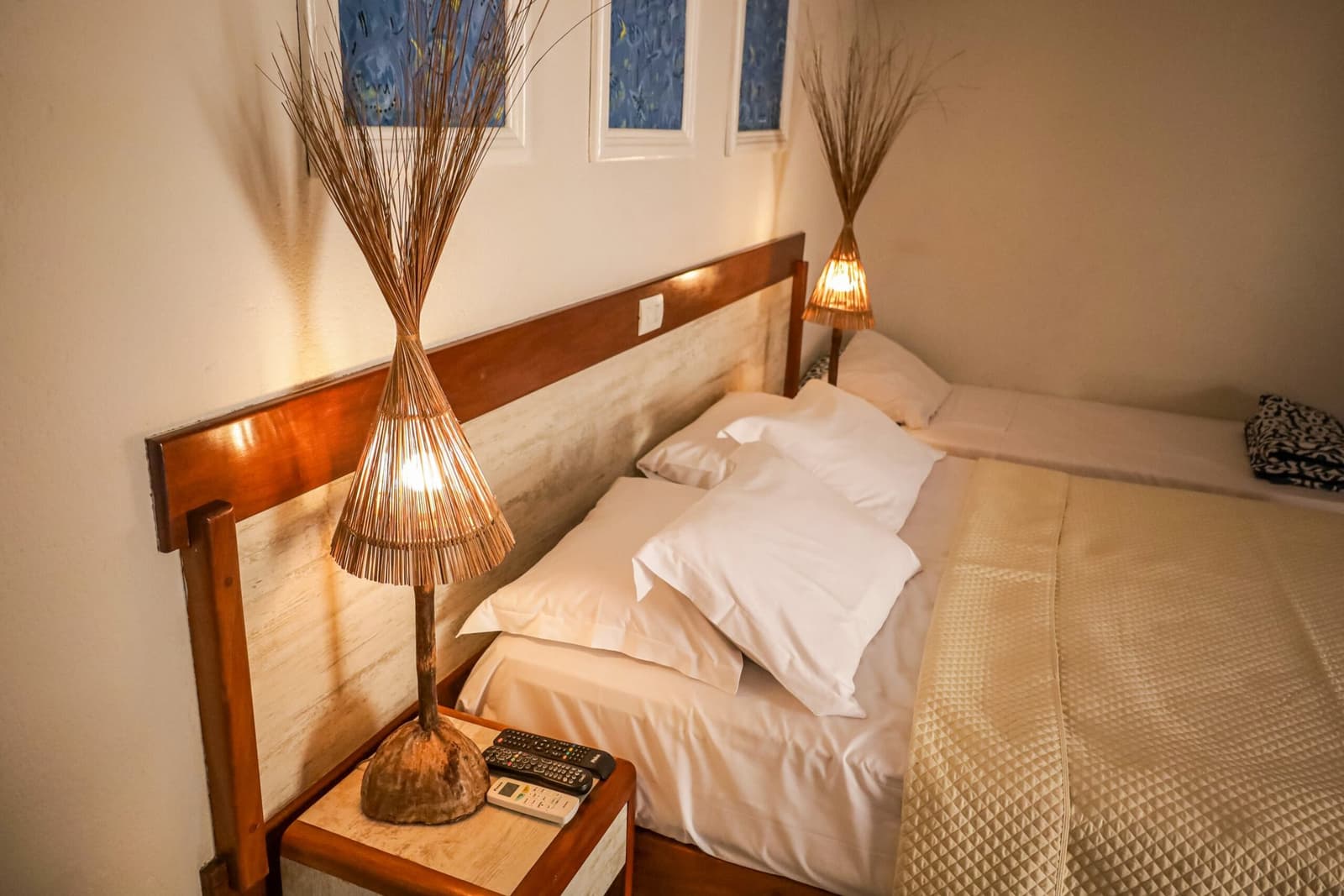 Klein, ruhig und genau richtig, nach einem langen Radeltag zählt das. Ein gemütliches, schlicht eingerichtetes Gästehausbzimmer auf der Fahrradroute Bahia Cocoa Route mit zwei Betten in weißer Bettwäsche, handgefertigten Rattanlampen auf einem Holznachttisch und blauen Zierfenstern über dem Kopfteil