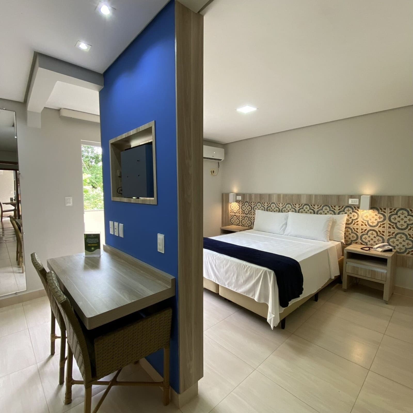 Nach dem Sattel kommt das Bett, und dieses macht seinen Job sehr gut. Helles Hotelzimmer mit Doppelbett, gemusterten Fliesen am Kopfteil, Holzschreibtisch, Rattanstühlen und einer blauen Akzentwand mit Wandmontage-TV