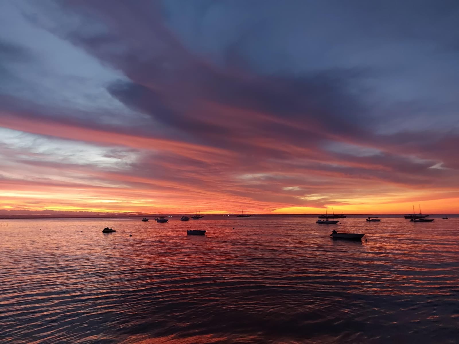 Am Abend gehört die Bucht euch ganz allein. Feuriger rot-oranger Sonnenuntergang über einer ruhigen Bucht mit silhouettierten Fischerbooten vor Anker, vom Ufer in Itacaré auf der Radroute durch Bahia