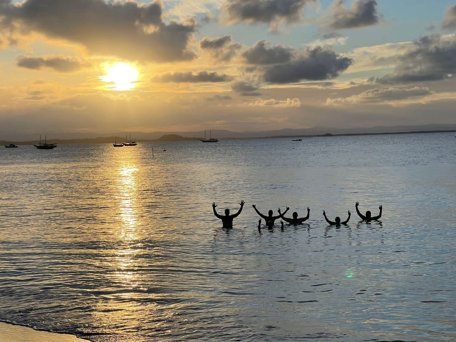 Arme hoch, Sonne unter, das Wasser sagt alles über diesen Abend. Fünf Personen als Silhouetten im Wasser mit erhobenen Armen bei einem goldenen Sonnenuntergang über einer ruhigen Bucht mit verankerten Segelbooten