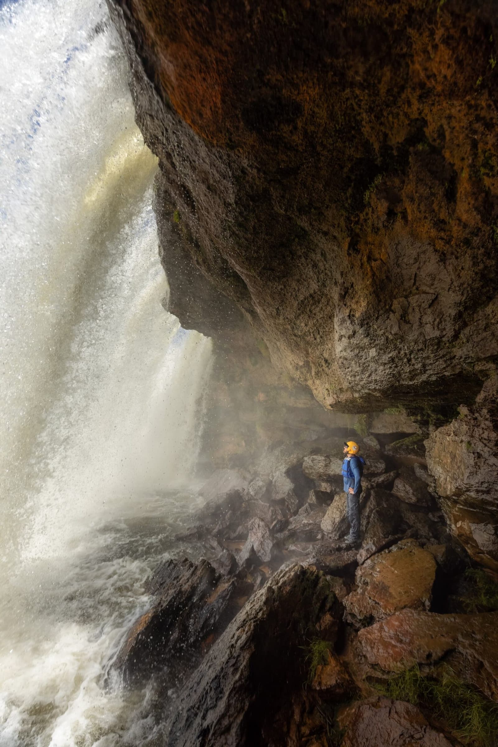 Mitten im Donner des Wasserfalls stehen, Cachoeira da Velha hält nichts zurück. Photo: Alexandre Socci Ein Wanderer in blauer Jacke und orangefarbenem Helm steht auf nassen Felsen unter einem gewaltigen Wasserfall-Überhang und blickt auf die donnernde Kaskade eines Jalapão-Trekkings