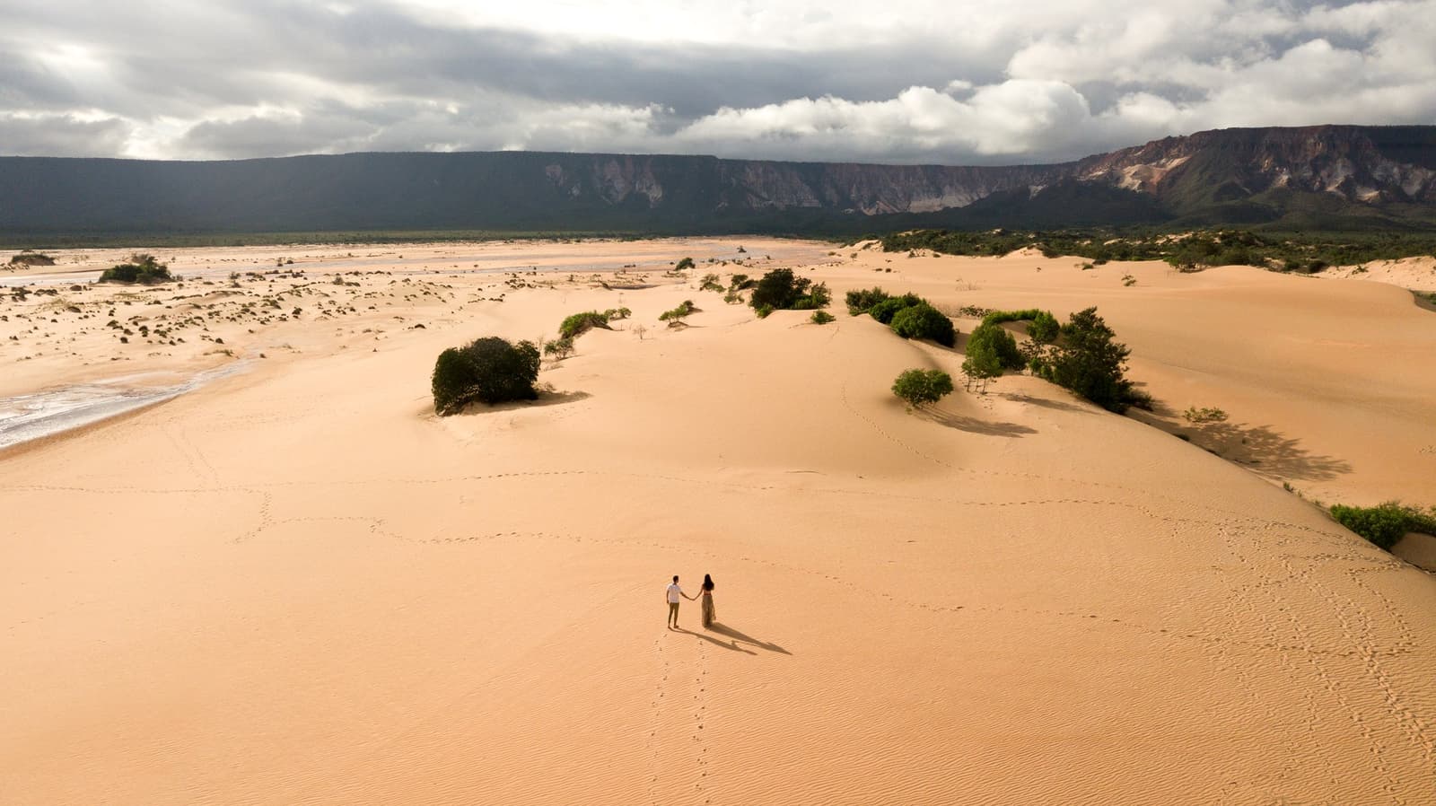 Zwei Menschen, ein Meer aus Sand, und Fußspuren, die genau dorthin führen, wo sie hin wollen. Photo: FG Trade Luftaufnahme eines Paares, das sich an den Händen hält und auf riesigen goldenen Sanddünen beim Jalapão-Trekking steht, mit grünen Sträuchern, einem Fluss und einem dramatischen Sandsteinfelsen unter bewölktem Himmel