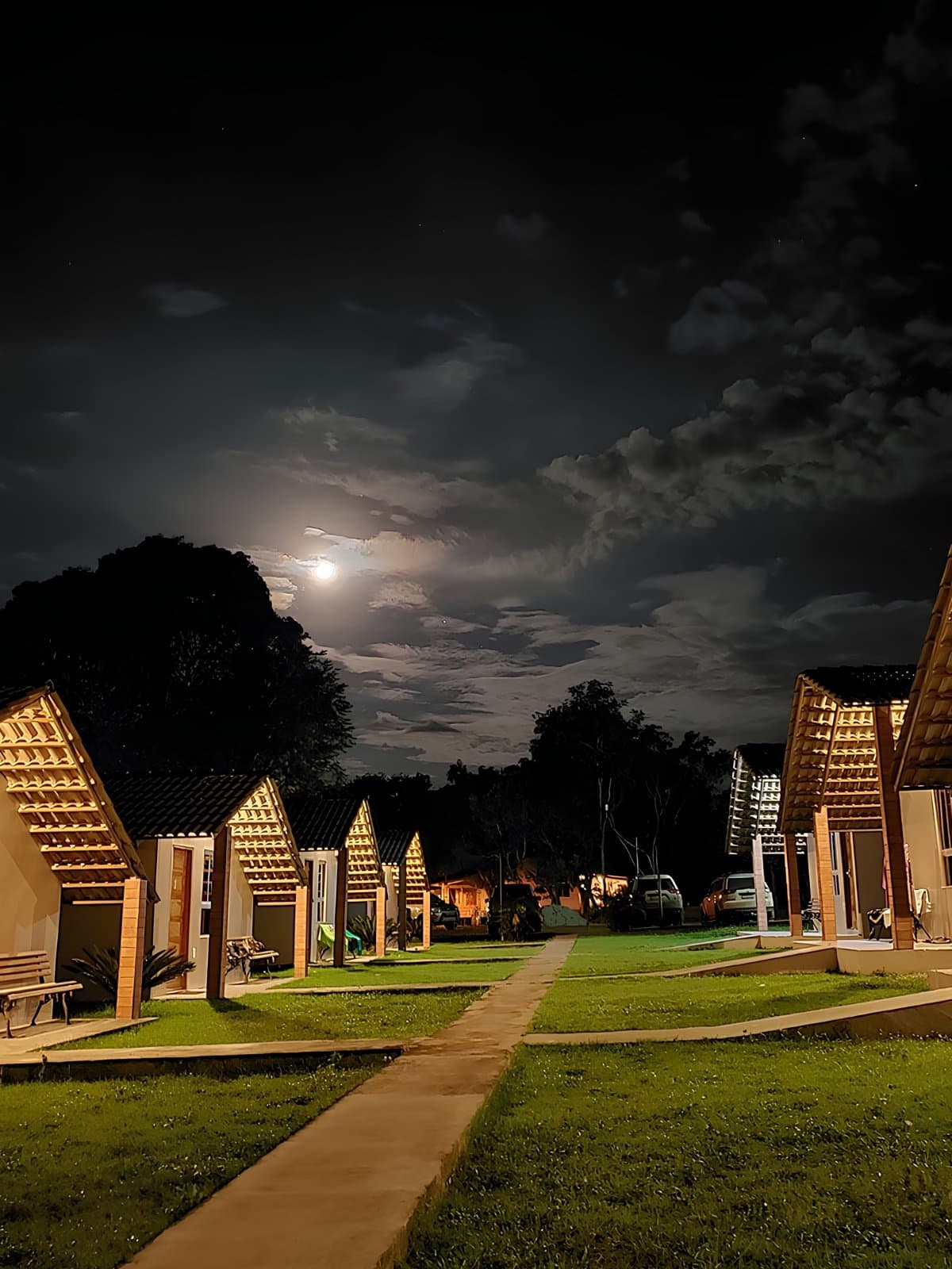 Müde Beine, gutes Abendlicht, strahlender Mond. Mehr braucht es nicht. Eine Reihe kleiner Holzdach-Chalets mit warmem Licht säumt einen Rasenweg bei Nacht, während ein Vollmond durch Wolken über den Bäumen bricht – ein ruhiger Abend im Basislager eines Jalapão-Trekking-Aufenthalts.