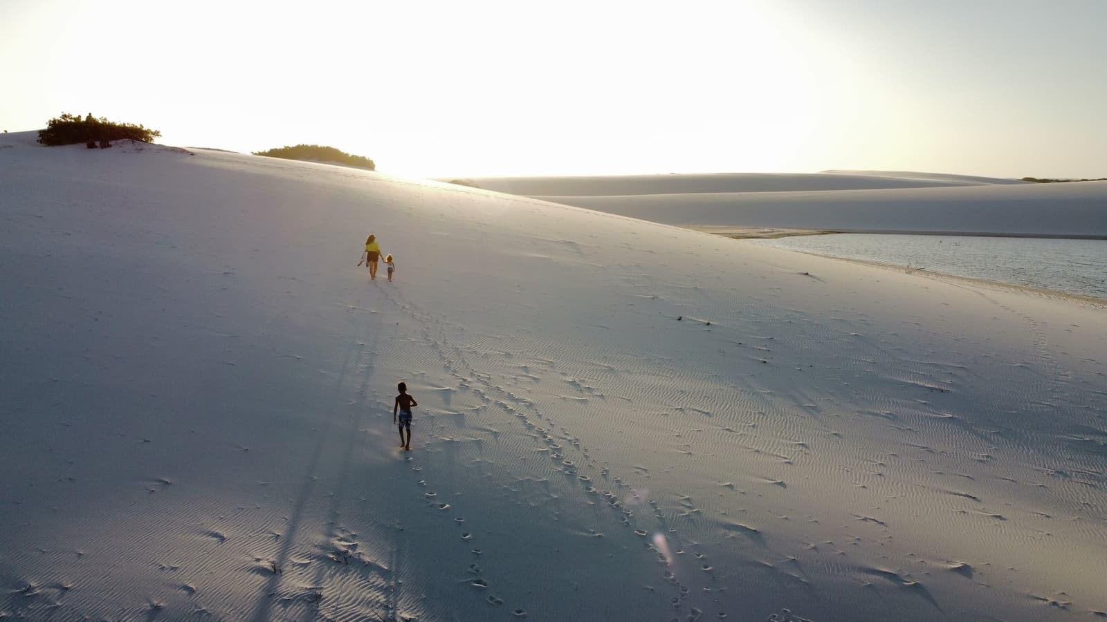 Gipfel ist Gipfel, egal wie klein die Beine sind. Eine erwachsene Person und zwei Kinder erklimmen eine riesige weiße Sanddüne neben einer stillen Lagune beim goldenen Stundenlicht im Lençóis Maranhenses