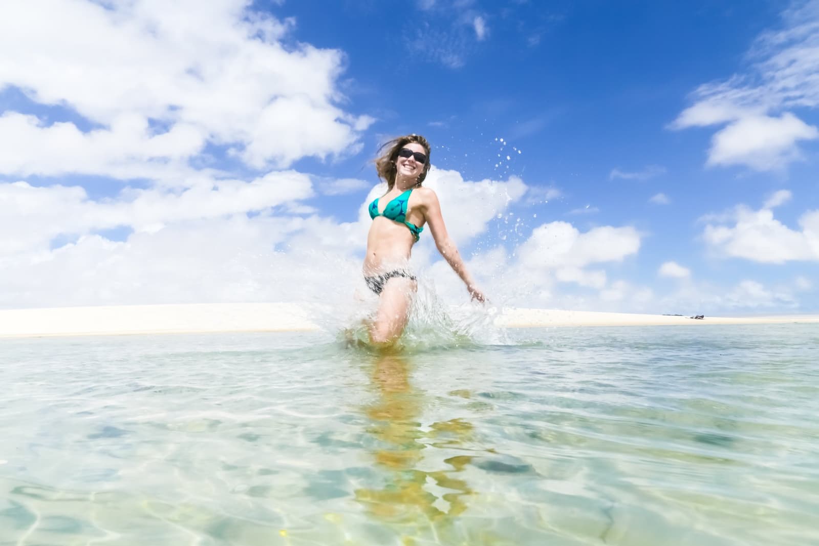 Kaltes Wasser, warmer Sand, keine Fragen mehr. Eine lächelnde Frau im türkisfarbenen Bikini planscht durch kristallklares, flaches Lagunenwasser vor weißen Sanddünen und strahlend blauem Himmel in Lençóis Maranhenses
