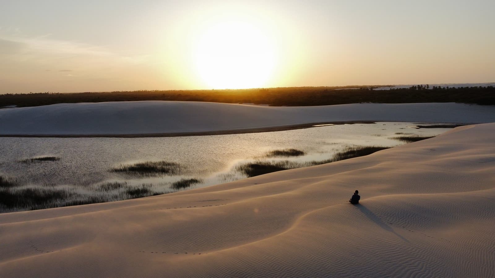 Dieser Moment gehört nur einer einzigen Person, und dem Horizont. Eine einsame Silhouette sitzt auf gewellten weißen Sanddünen und blickt bei goldenem Sonnenuntergang auf eine flache, grasgesäumte Lagune — ein unvergesslicher Moment der Lençóis Maranhenses Rundreise