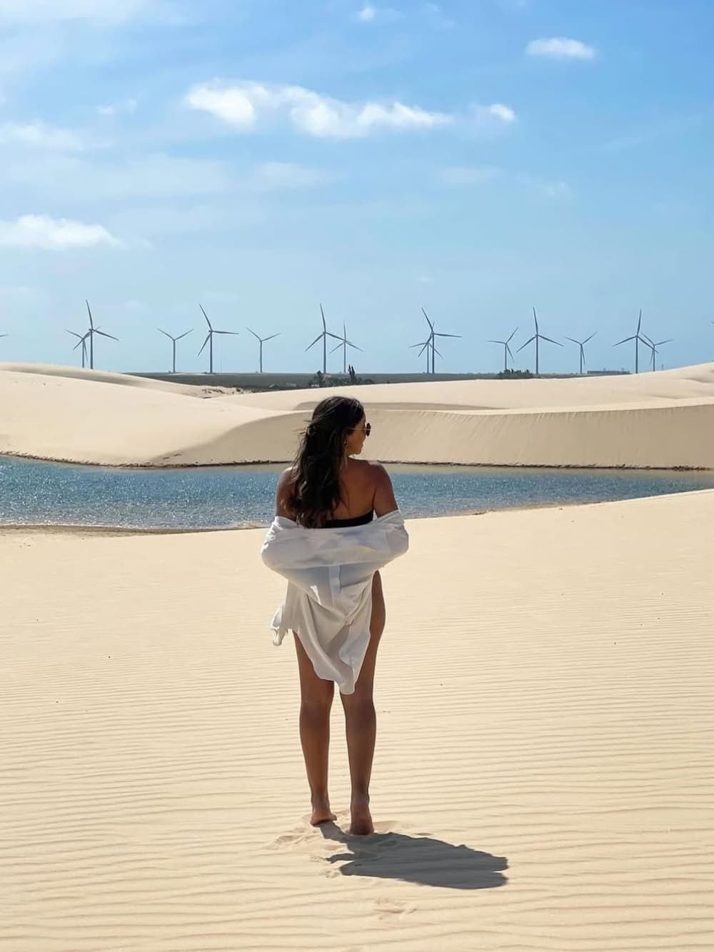 Sand, Wasser und Windräder, hier sieht die Natur die Moderne einfach kommen. Eine Frau in weißem Hemd steht barfuß auf gerippten weißen Sanddünen und blickt auf eine glitzernde blaue Lagune, während sich eine Reihe Windräder am Horizont erstreckt — ein beeindruckendes Bild der vollständigen Reiseroute durch Lençóis Maranhenses