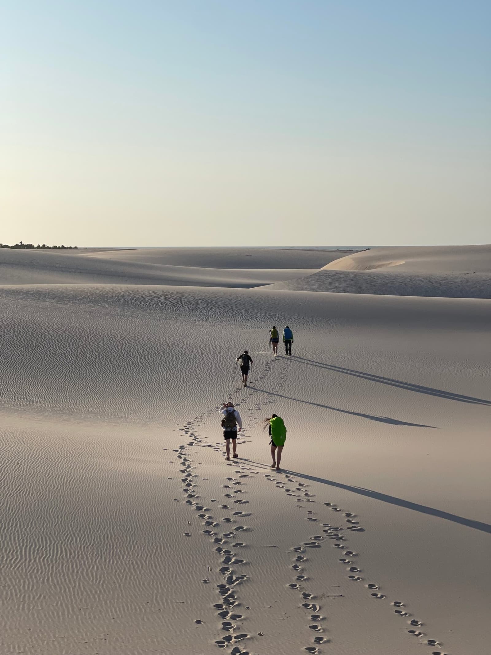 Vier Wanderer, unzählige Schritte, und das Gefühl, ganz allein auf der Welt zu sein. Vier Wanderer mit Rucksäcken überqueren bei einer Lençóis-Maranhenses-Wanderung weite weiße Sanddünen, ihre Fußspuren ziehen sich weit hinter ihnen her unter einem hellen Morgenhimmel