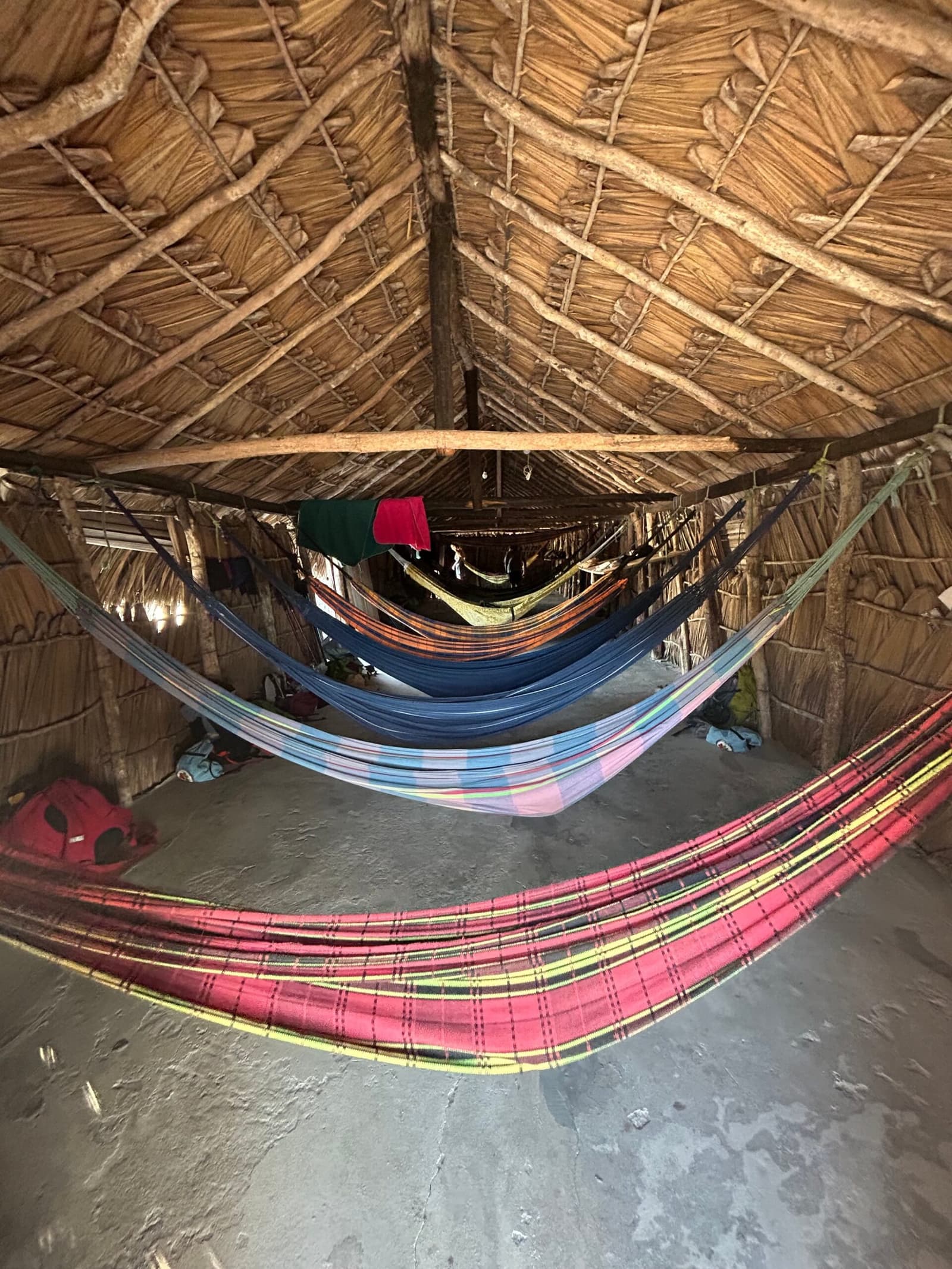 Fünf Farben, fünf Hängematten, ein sehr guter Schlaf. Mehrere bunte Hängematten in einer rustikalen Unterkunft mit Strohdach beim Lençóis-Maranhenses-Trekking, mit Reisegepäck auf dem Lehmboden und warmem Licht durch die geflochtenen Wände