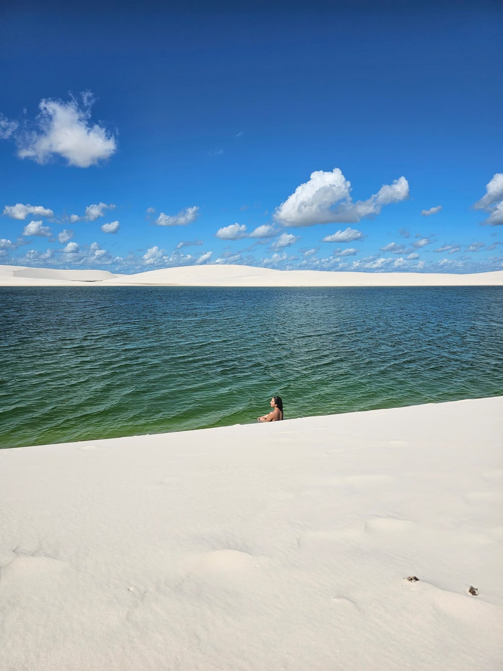 Smaragdgrünes Wasser mitten in einer Dünenlandschaft. Brasilien macht manchmal keinen Sinn, und das ist gut so. Eine Frau steht bis zur Brust in einer klaren grün-blauen Süßwasserlagune der Lençóis Maranhenses, dahinter riesige weiße Dünen unter einem strahlend blauen Himmel