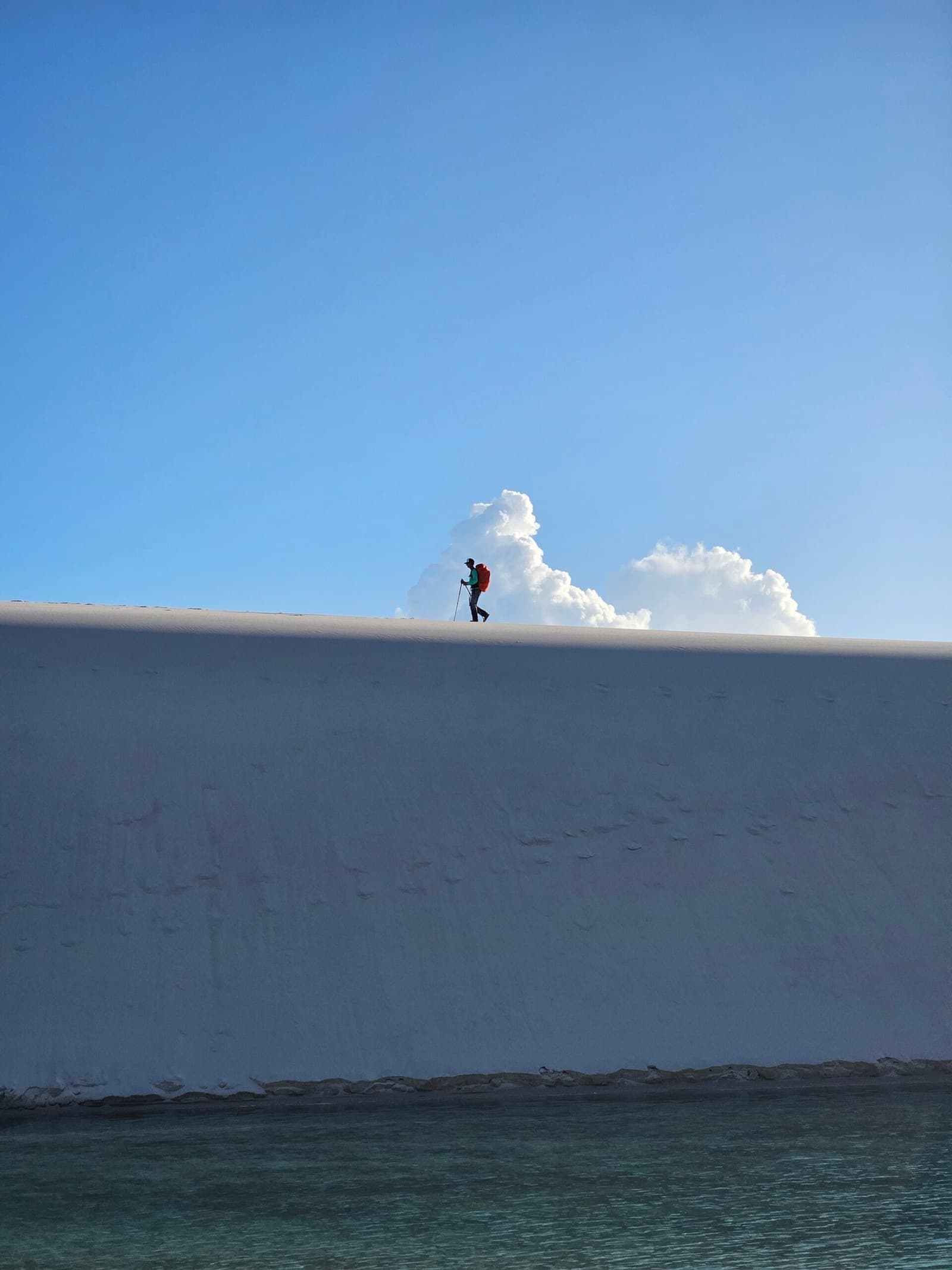 Eine Silhouette, eine Düne, weiter Himmel. Mehr braucht es nicht. Ein einsamer Wanderer mit rotem Rucksack und Trekkingstock läuft auf dem scharfen Kamm einer hohen weißen Sanddüne bei der Lençóis-Maranhenses-Wanderung, darunter eine türkisfarbene Lagune und aufgetürmte Wolken dahinter