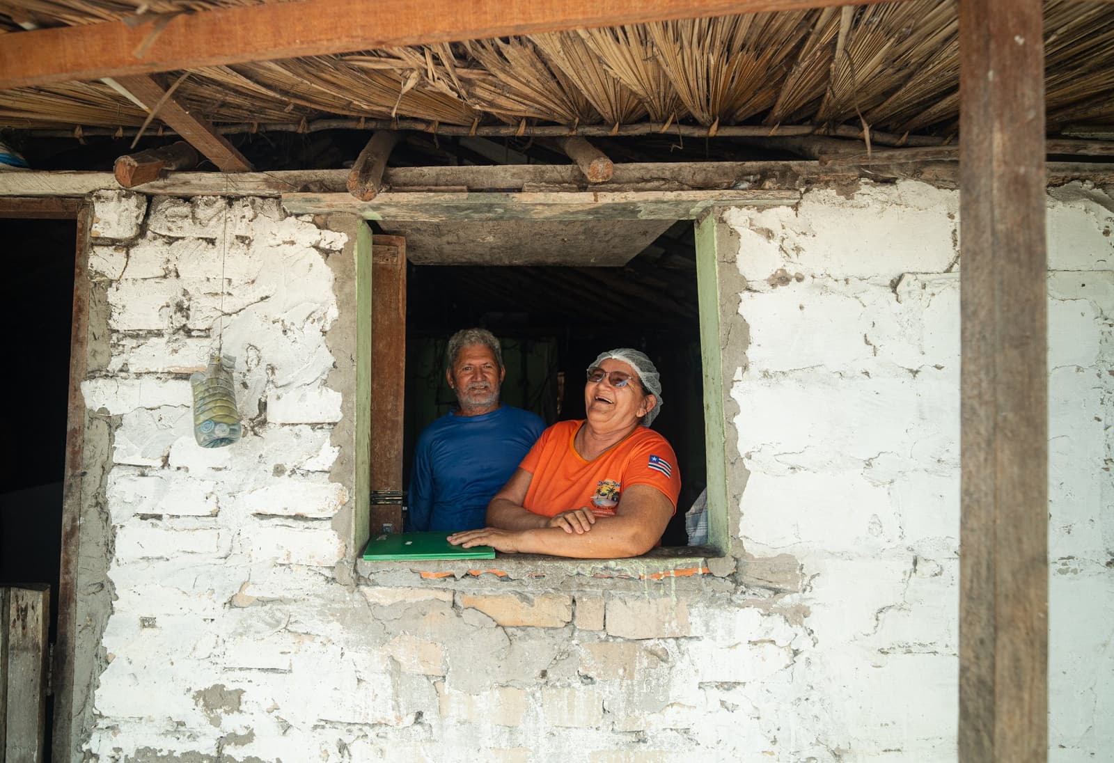 Un sourire à la fenêtre, et vous vous sentez déjà invités. Un homme en t-shirt bleu et une femme en t-shirt orange rient ensemble depuis la fenêtre d'une maison en brique blanchie à la chaux avec un toit de chaume, famille d'accueil rencontrée lors d'un trek dans les Lençóis Maranhenses.