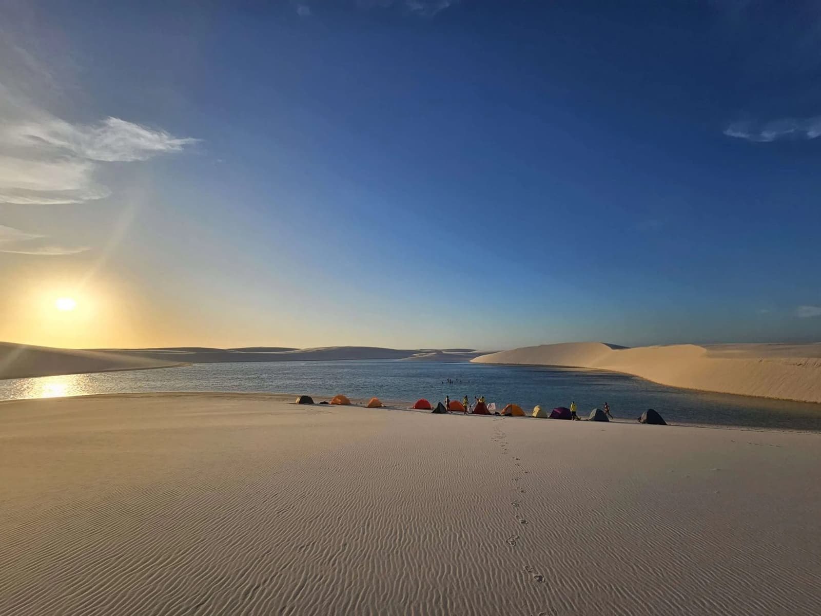 Ce soir, le campement s'installe là où les dunes rencontrent l'eau. Difficile de faire mieux. Des tentes colorées dressées au bord d'une lagune d'eau douce au coucher du soleil lors d'un trek dans les Lençóis Maranhenses, avec des dunes de sable blanc ondulées s'étendant à l'horizon sous un ciel bleu intense