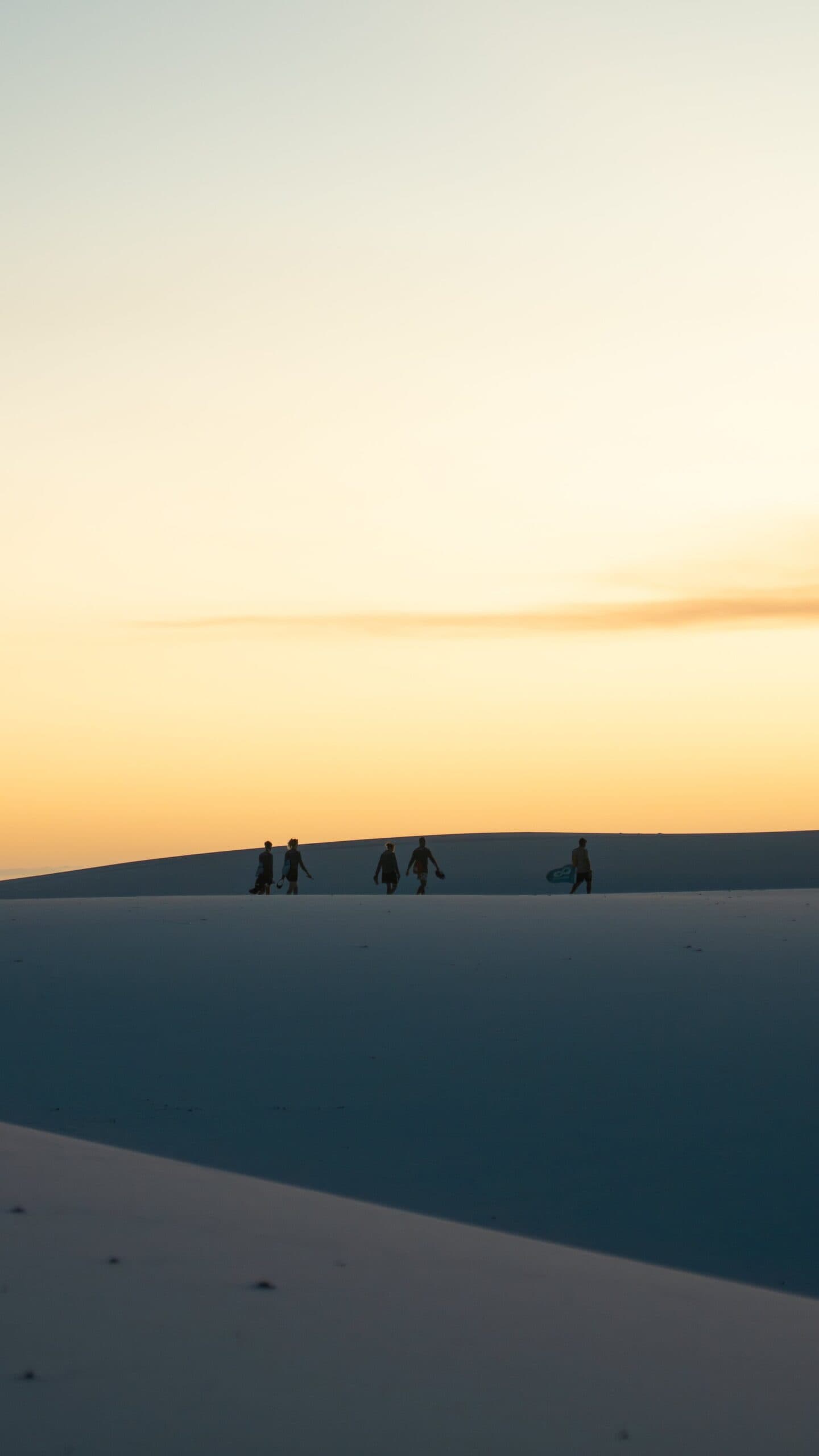 Lumière de fin de journée, planche sous le bras, et une dune qui n'en finit pas. Cinq silhouettes, dont une portant un sandboard, marchent sur la crête d'une dune de sable blanc au coucher du soleil lors d'un trek dans les Lençóis Maranhenses, sur fond de ciel doré