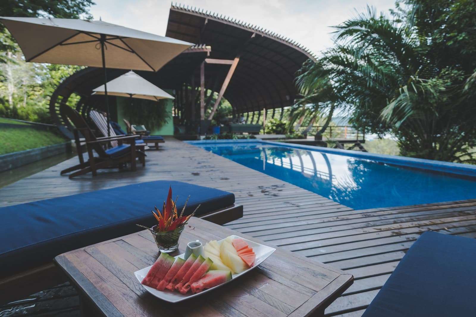 Frische Früchte, kühles Wasser und der Regenwald direkt nebenan. Mehr braucht der Nachmittag nicht. Ein Teller mit aufgeschnittener Wassermelone und Ananas auf einem Liegestuhl-Tisch am Pool des Mirante do Gavião, mit einem geschwungenen Holzpavillon und üppigem tropischem Grün im Hintergrund