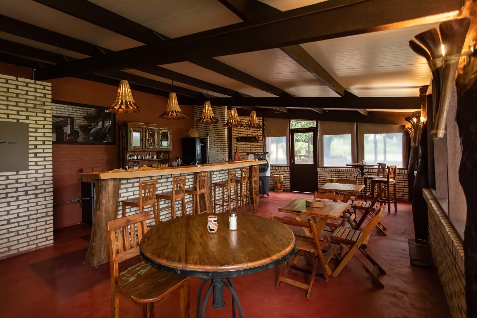 Warm wood, woven lampshades, and cold drinks waiting after a long day on the floodplain. Rustic dining room and bar of a southern pantanal safari lodge, featuring wooden round tables, high stools along a brick-faced counter, handcrafted pendant lights, and large windows overlooking green vegetation outside
