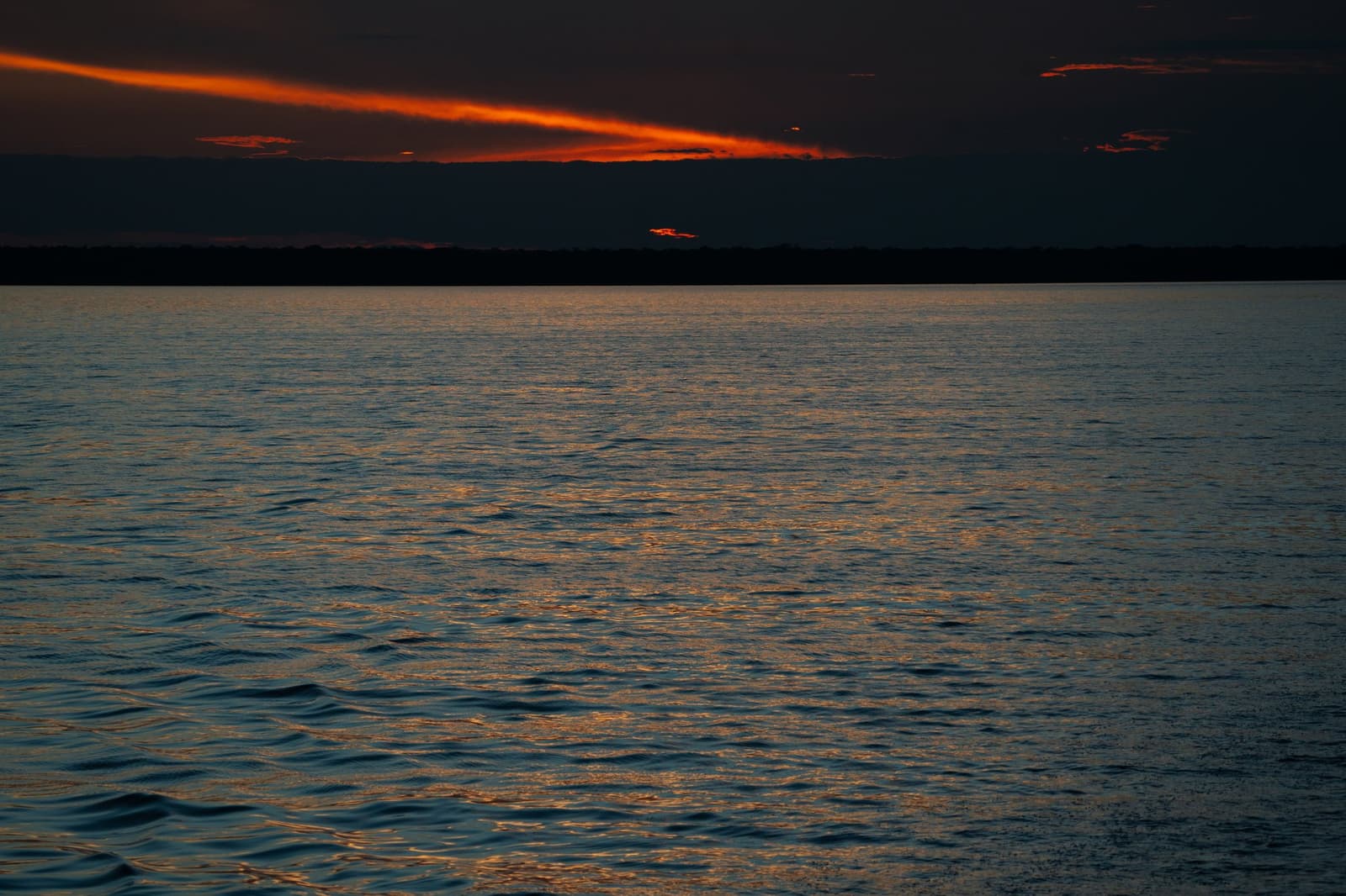 Das letzte Licht des Tages, und der Fluss lässt nichts davon los. Tiefrot und orange leuchtende Wolkenstreifen spiegeln sich über einem breiten, dunklen Fluss in der Dämmerung auf der Tucano Amazon River Cruise, mit einem waldgesäumten Horizont vor fast schwarzem Himmel